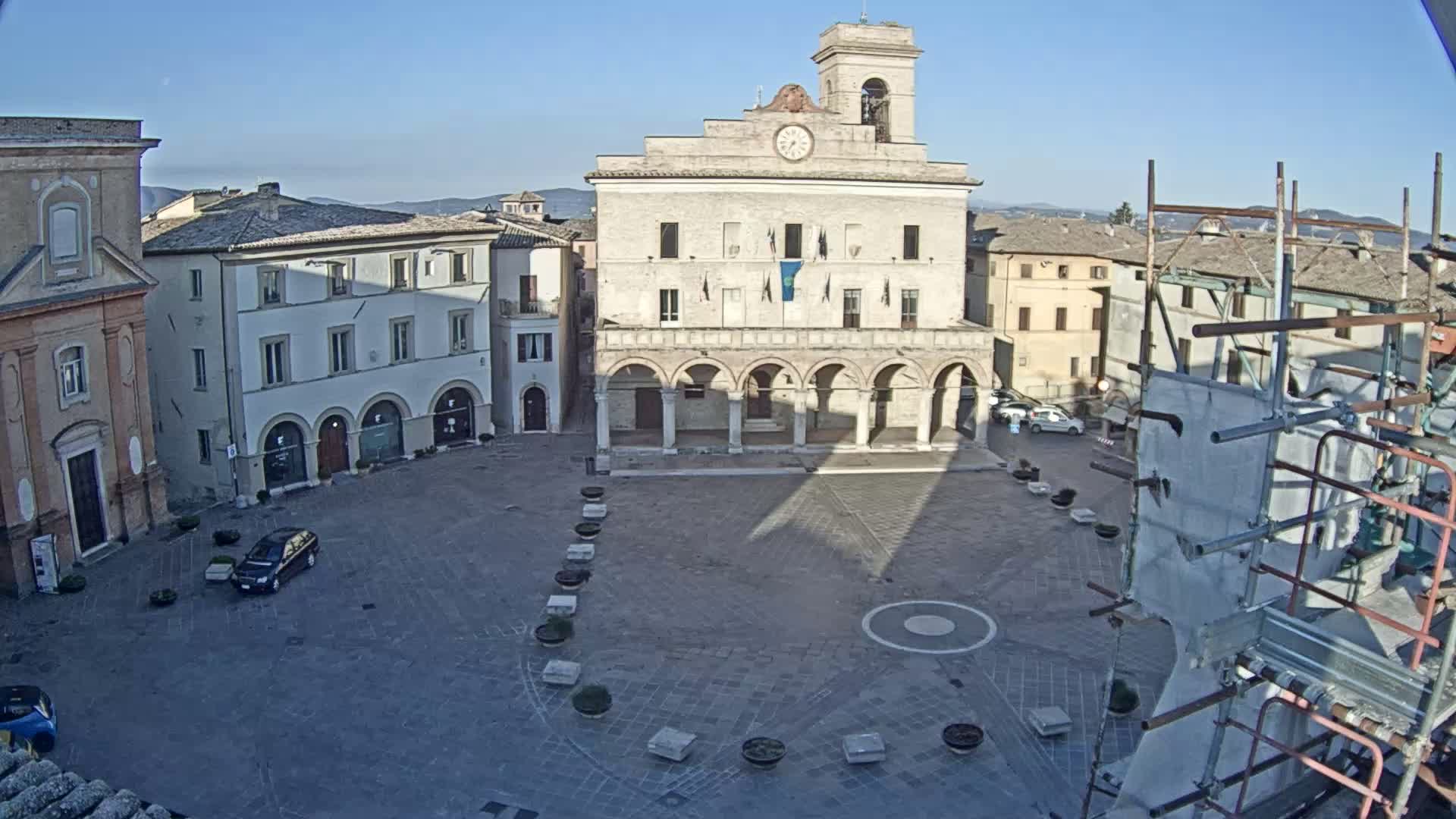 A town square with a large building featuring arched entryways is seen on a sunny day, with scaffolding and a few cars visible.