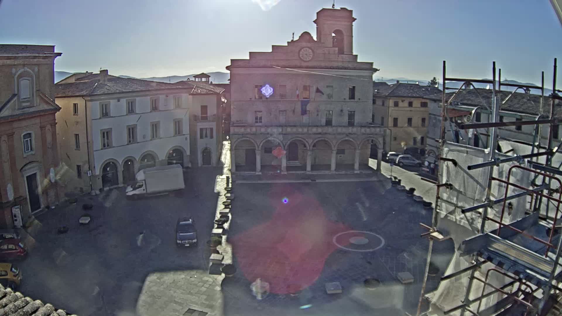A sunny, high-angle view of a town square dominated by a large, pale stone building with arched entryways, surrounded by other buildings and some vehicles, with scaffolding visible in the foreground.
