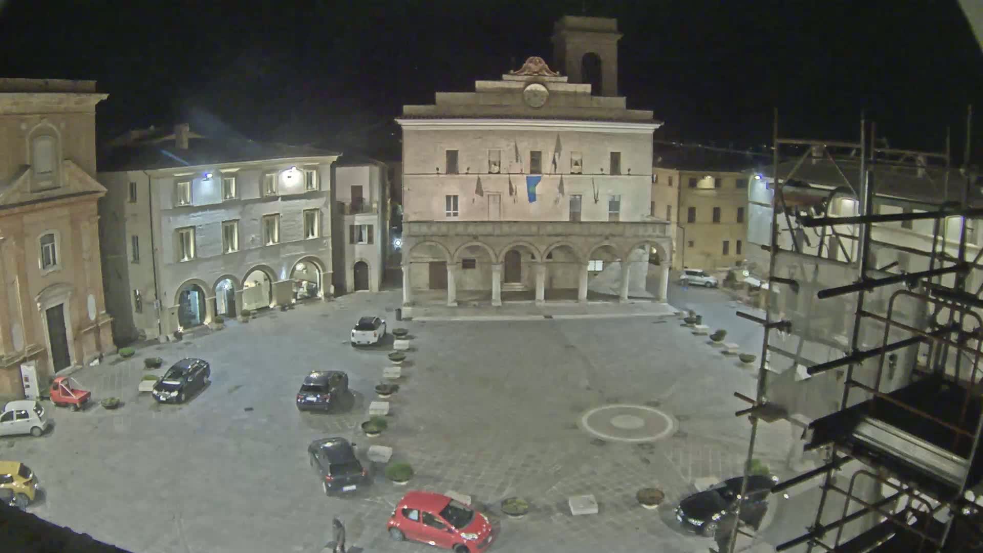 An empty town square at night, illuminated, with several cars parked around its perimeter and scaffolding on a building in the background.