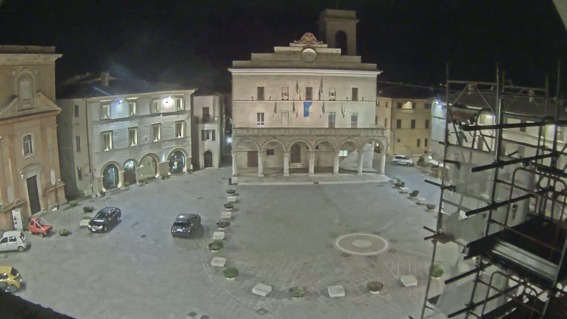 A nighttime view of a town square with several buildings, including a large building with arched entryways, a few parked cars, and scaffolding.