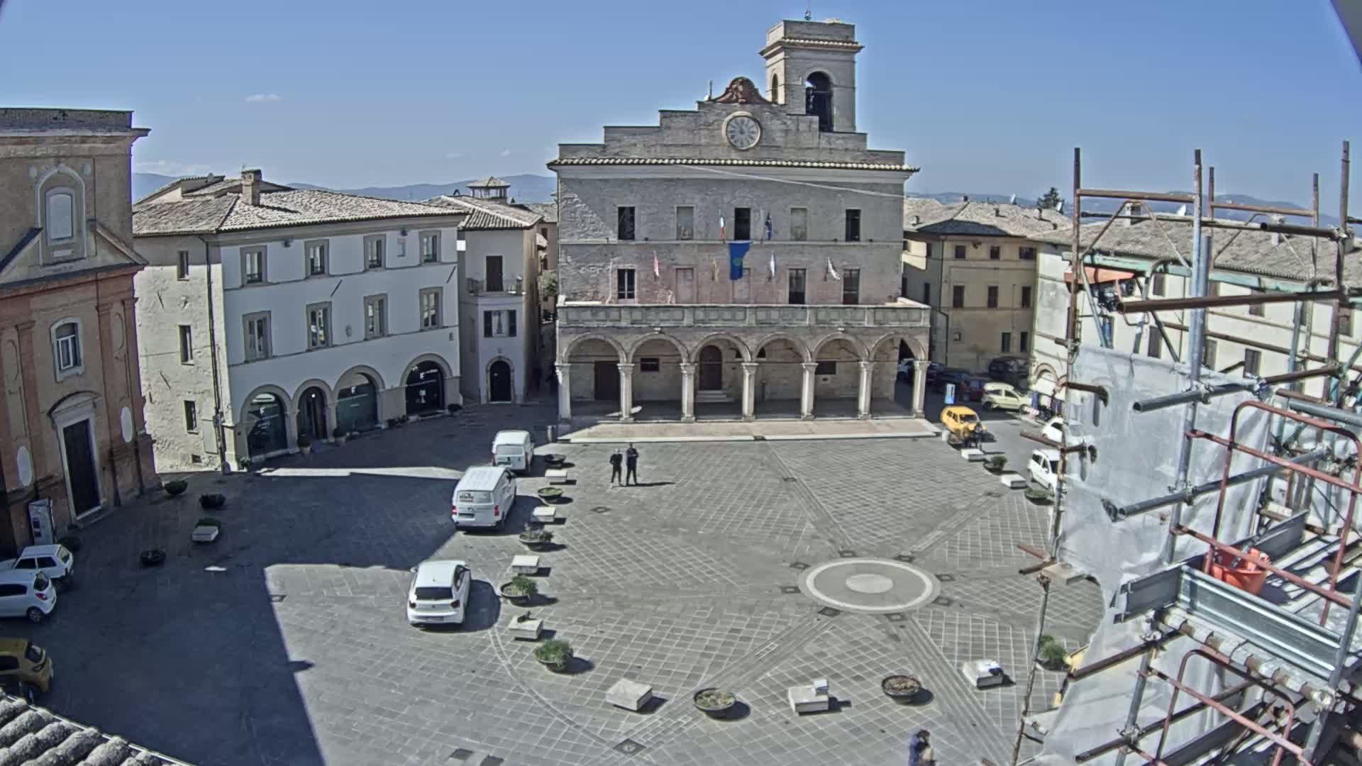 A town square with a large stone building, several parked cars, and scaffolding on a sunny day.