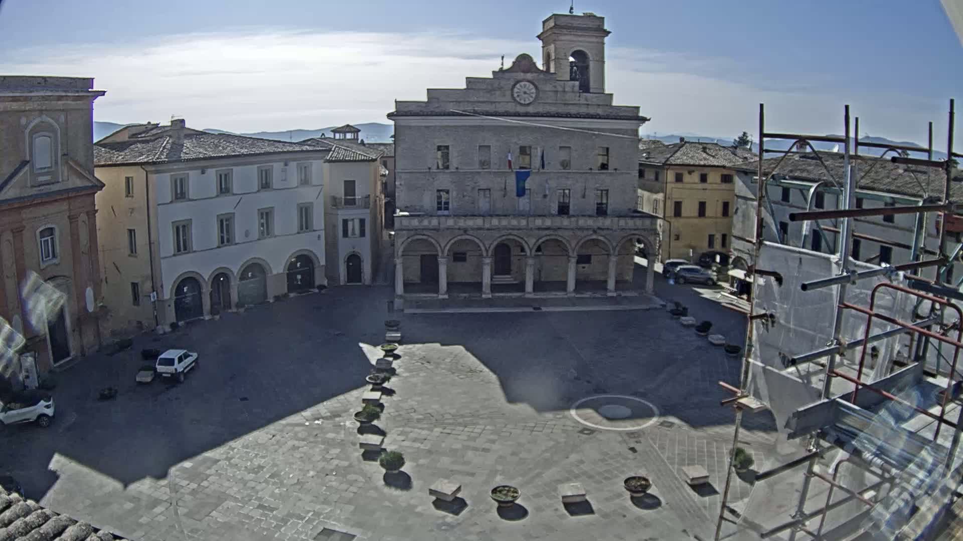 A sunny town square is seen from above, centered on a large building with arched colonnades, surrounded by other buildings, a few cars, and scaffolding.