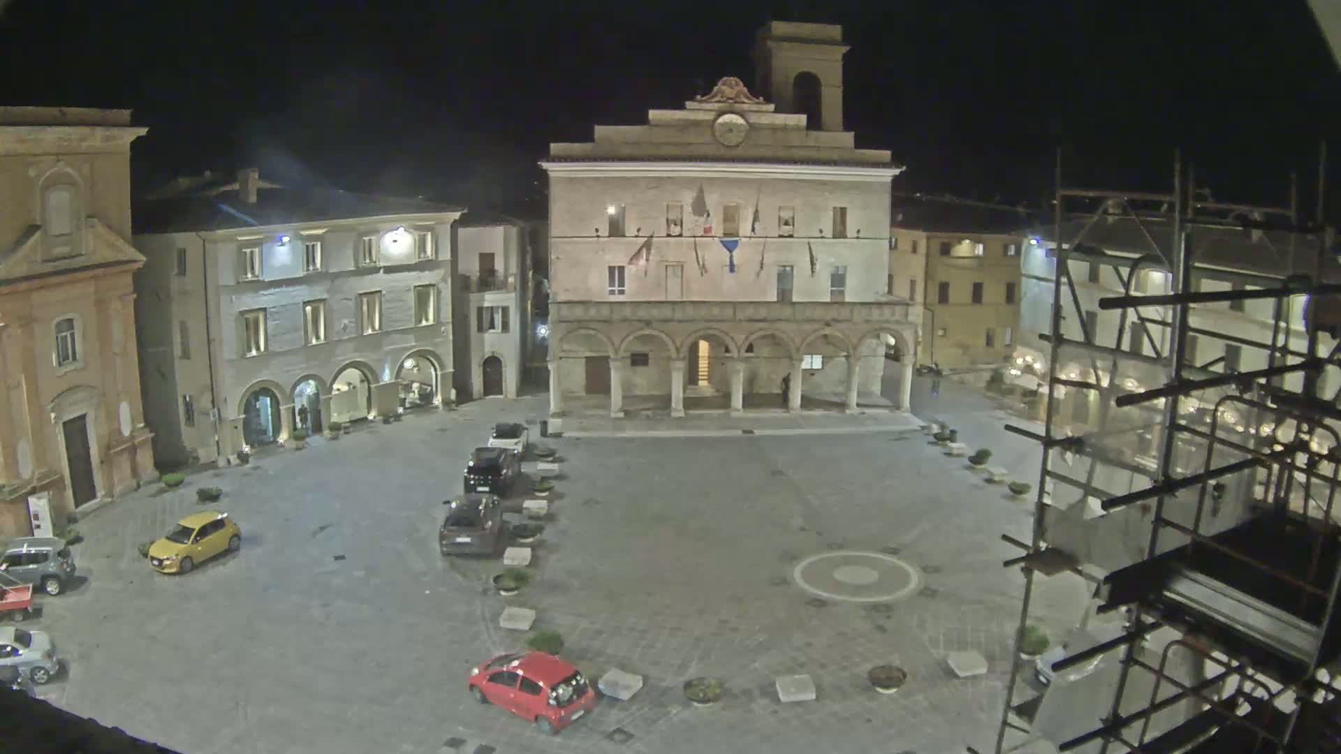 A nighttime view of a town square, featuring a large building with arched entryways, several cars parked in the square, and scaffolding surrounding a building under construction.