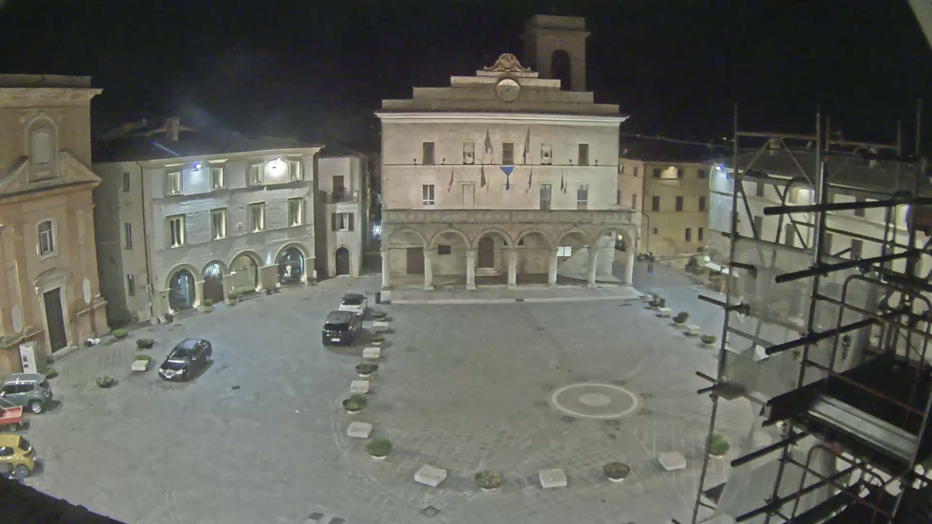A town square at night, illuminated, features several buildings including a large structure with arched entryways, and a few cars are parked in the mostly empty square.