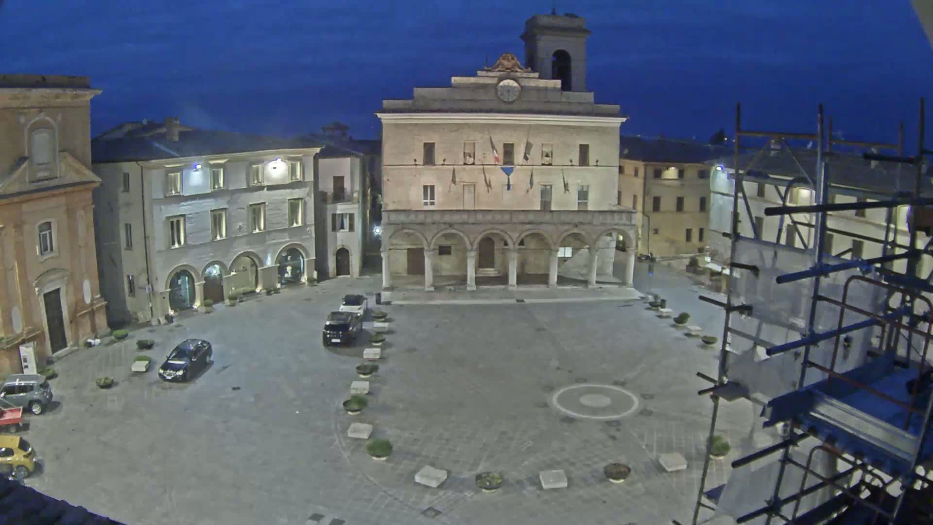 A town square at night, under a dark blue sky, features a large building with arched entryways, several cars parked in the square, and scaffolding to the right.