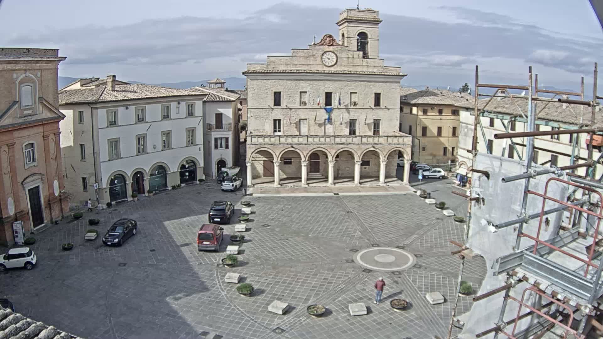 A mostly empty town square with a large, arched building in the center is visible under a partly cloudy sky, with scaffolding surrounding a portion of one building.