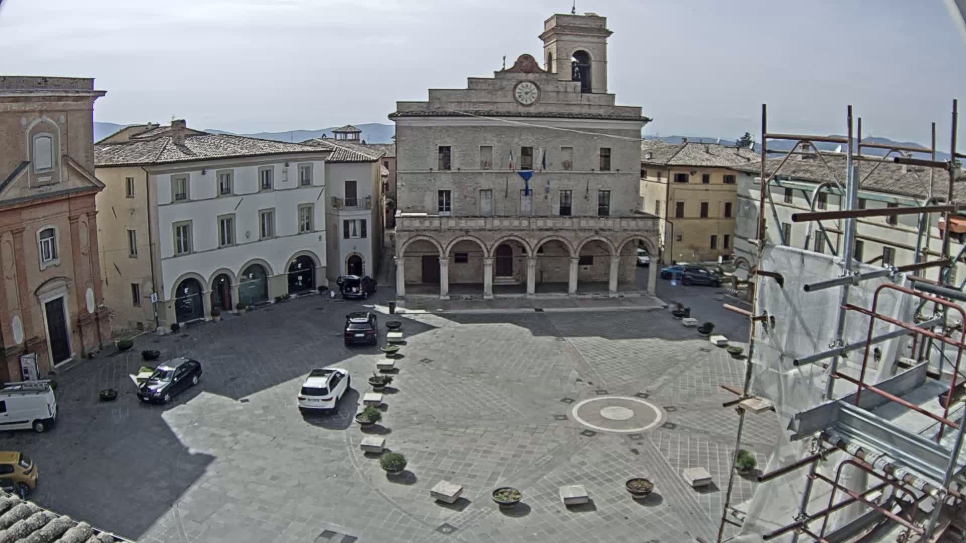 A town square, under a mostly sunny sky, is centered around a large building with arched entryways, and surrounded by other buildings and a few parked cars.