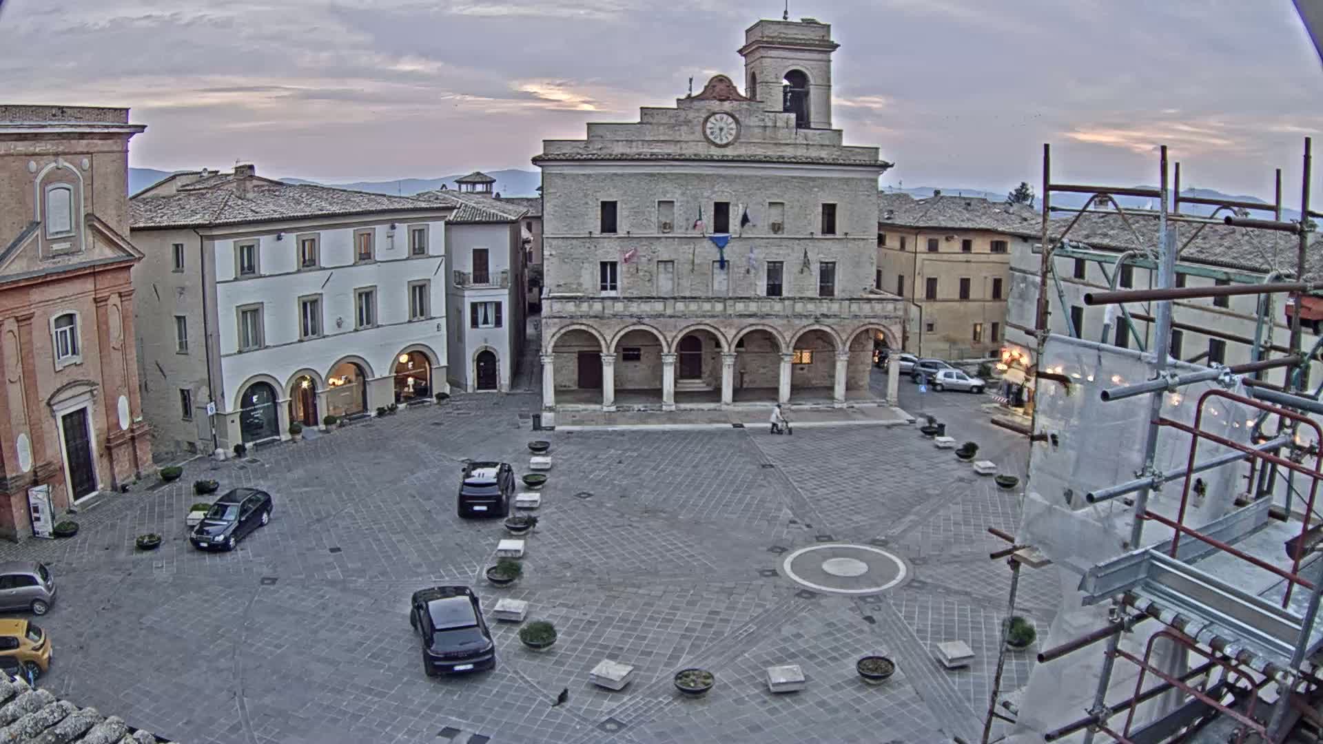 A town square with several cars parked in it, surrounded by historical buildings under an overcast sky.