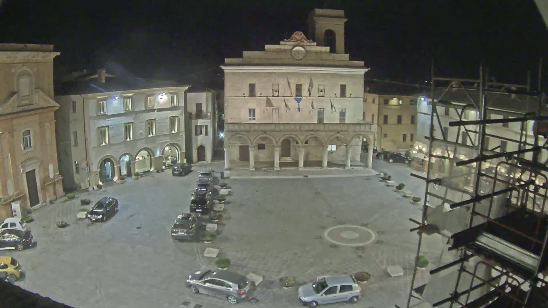 A nighttime view of a town square with several parked cars,  surrounded by historic buildings, one of which is a large, arcaded structure.