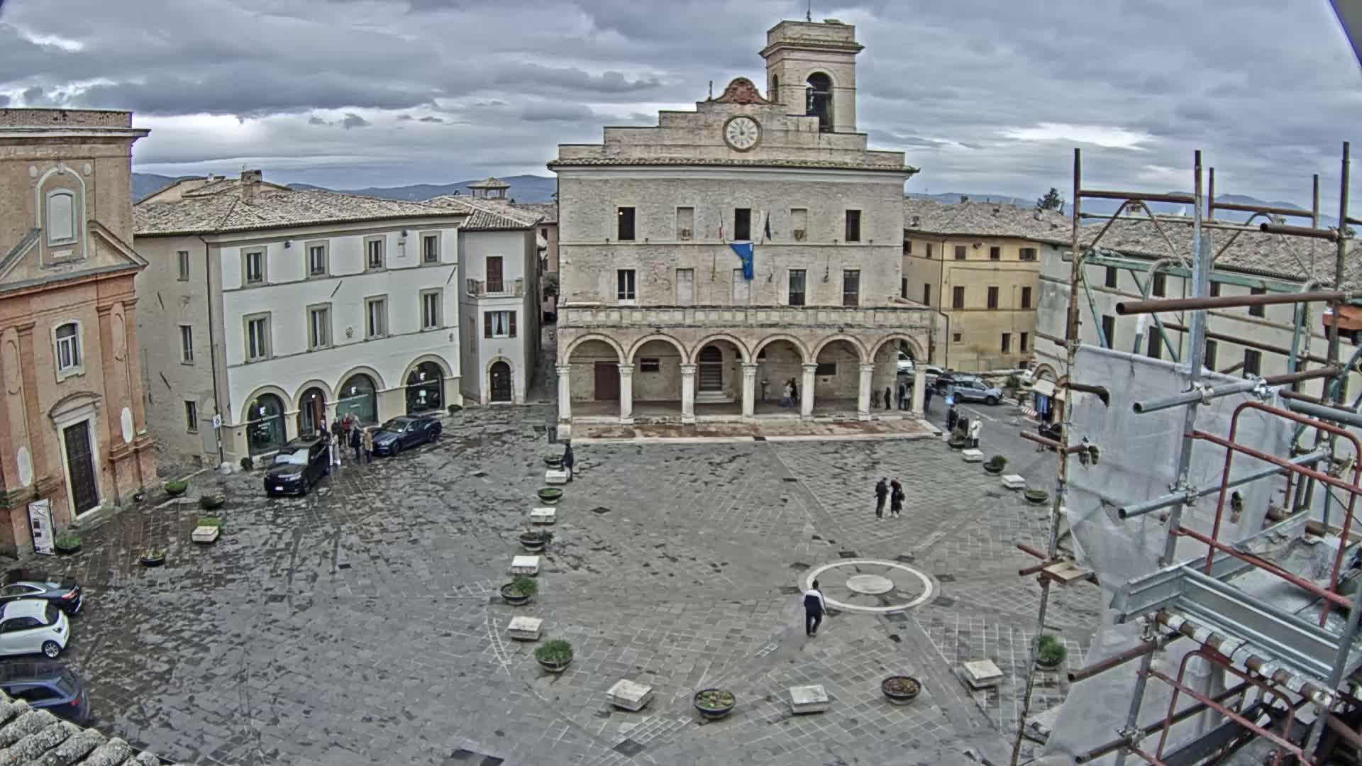 A town square with a large stone building, surrounded by other buildings, under an overcast sky with light rain.