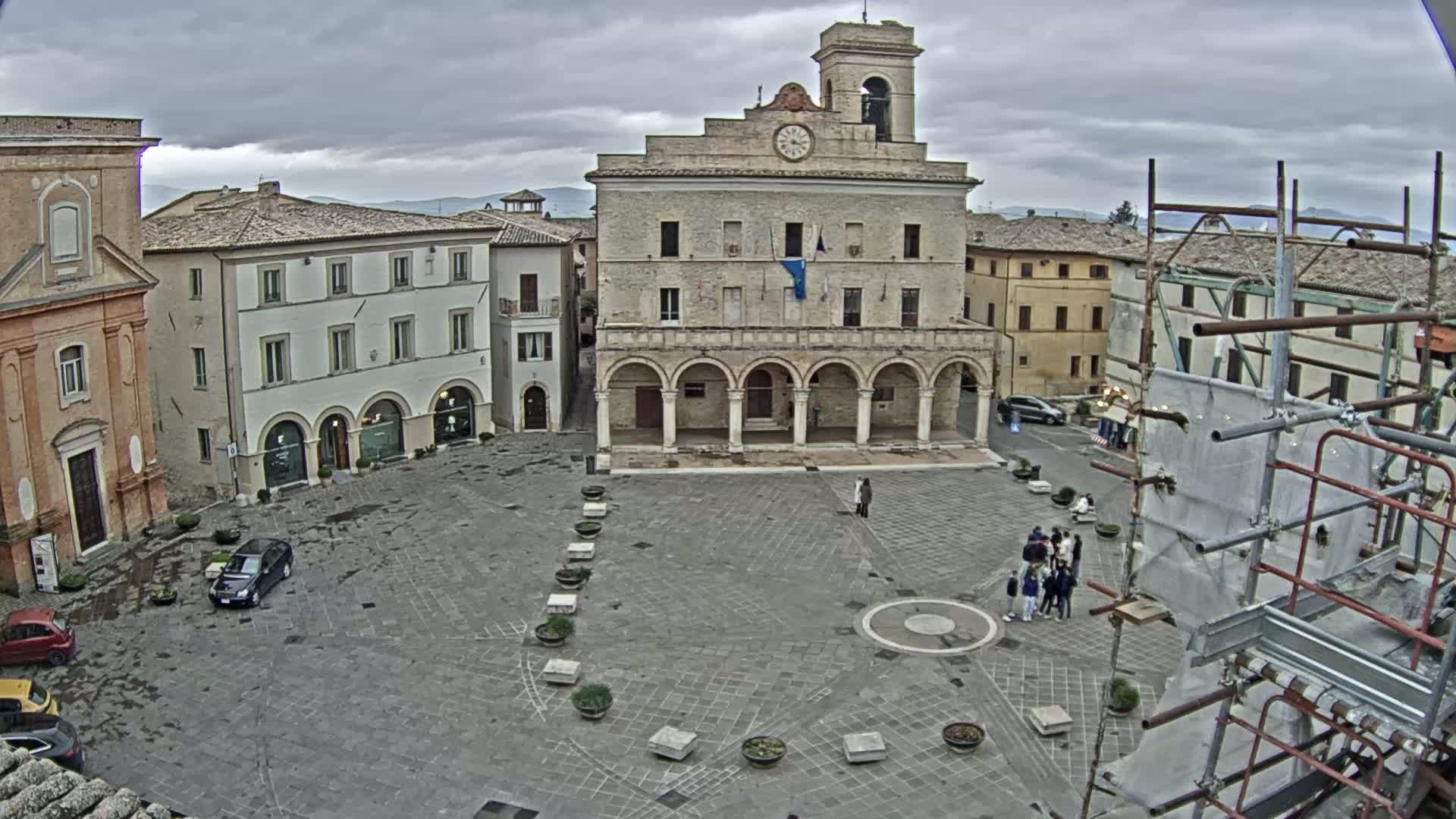 A town square with a large stone building, several other buildings, a few cars, and some people standing in a group, under an overcast sky.