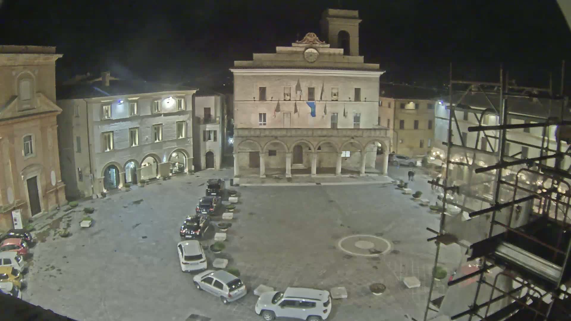 A nighttime aerial view of a town square shows several cars parked along one side, an ornate building with arched entryways in the center, and other buildings surrounding the square under dark, clear skies.