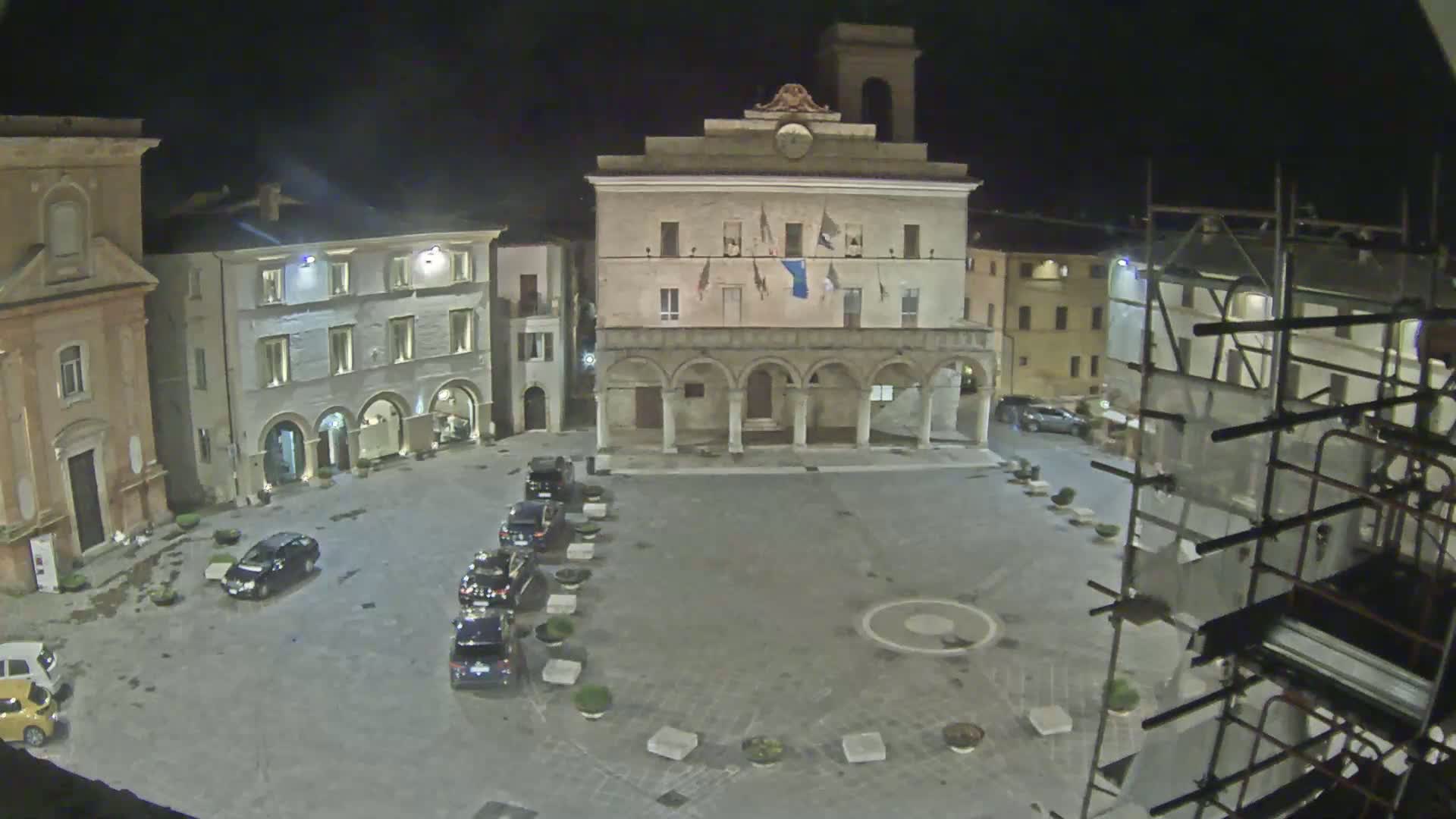 A town square at night is shown, with several cars parked along one side and a large building with arched entryways at the center, under dark, clear skies.