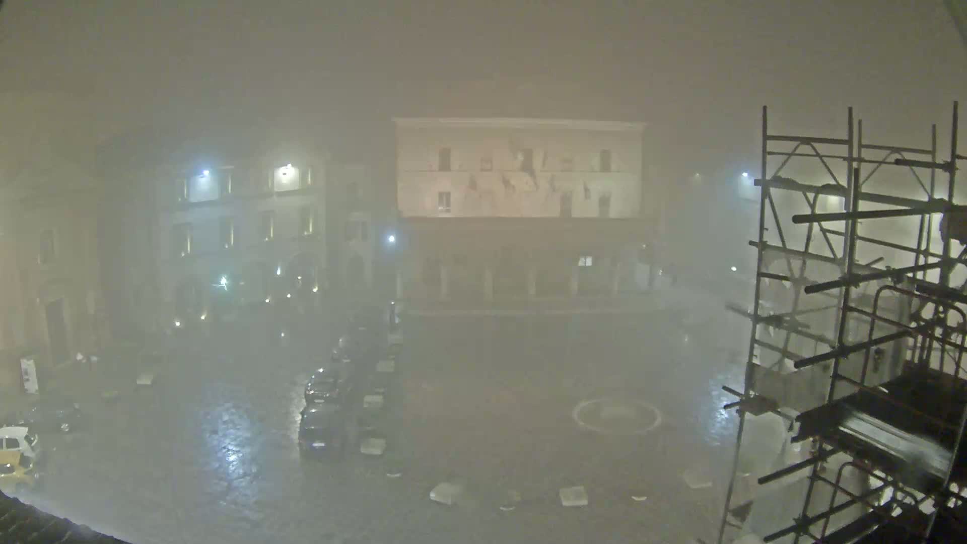 A foggy, nighttime view of a town square with several buildings, parked cars, and scaffolding.
