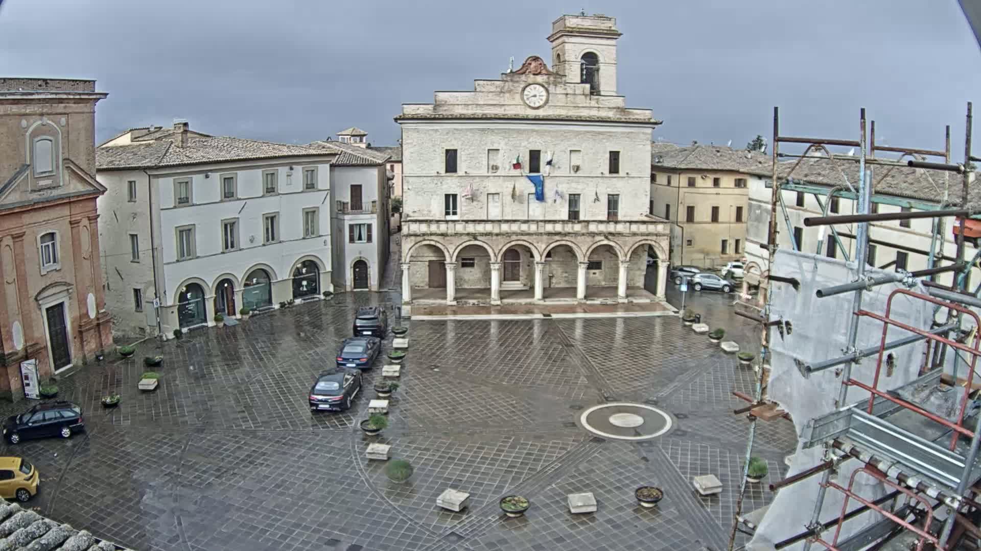 A wet, paved town square is centered around a large, light-colored building with arched entryways, surrounded by other buildings under a cloudy sky.