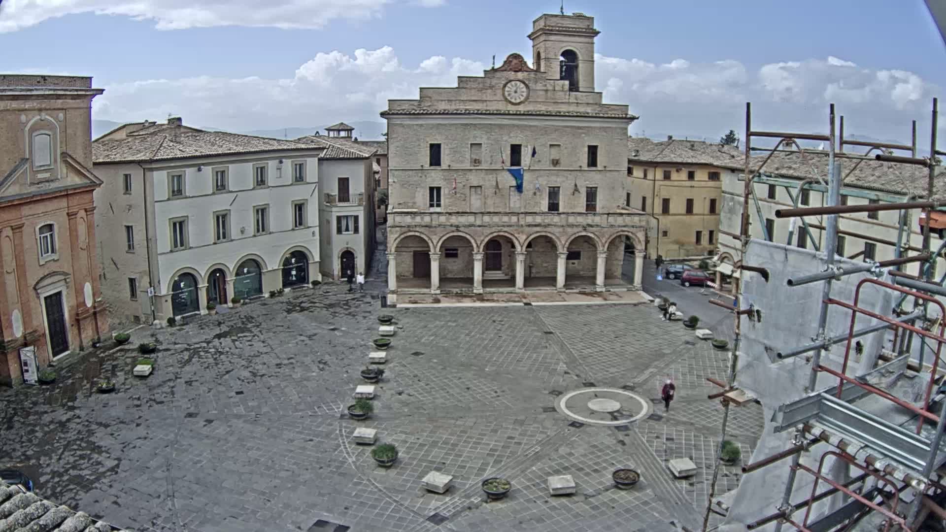 A large, stone plaza is surrounded by historic buildings under a partly cloudy sky, with scaffolding visible on the right.