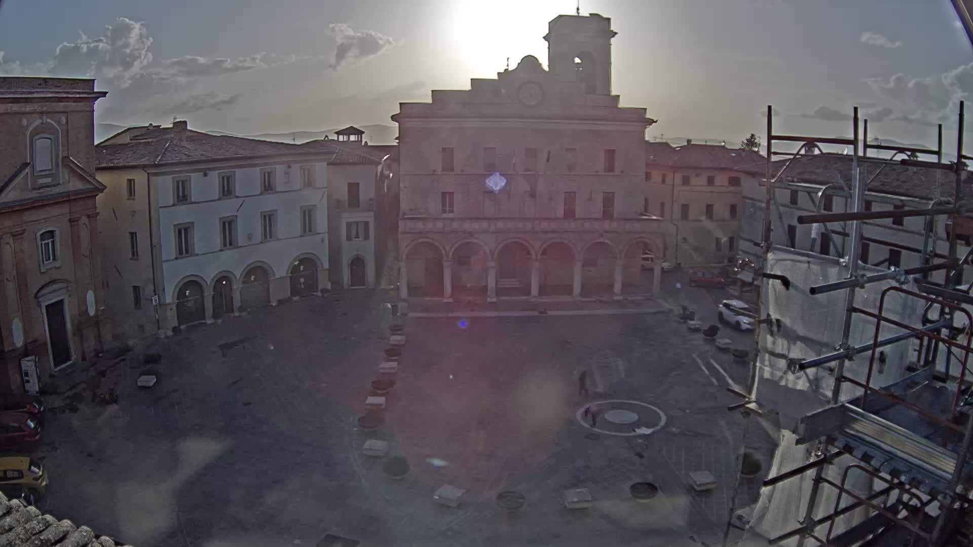 A sunlit town square, partially surrounded by old buildings, features a large central building with arched entryways and a clock tower, under partly cloudy skies.