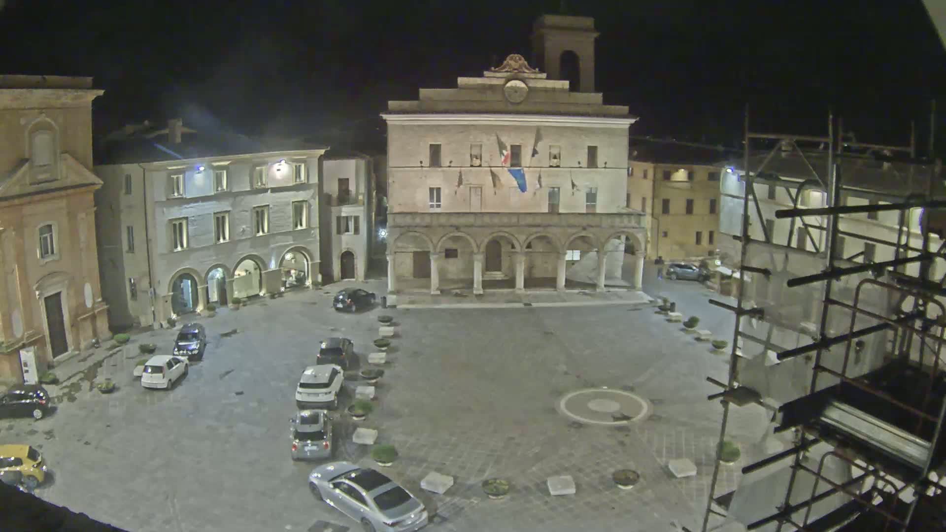 A nighttime view of a town square with several cars parked, dominated by a large building with arched entrances and a bell tower.