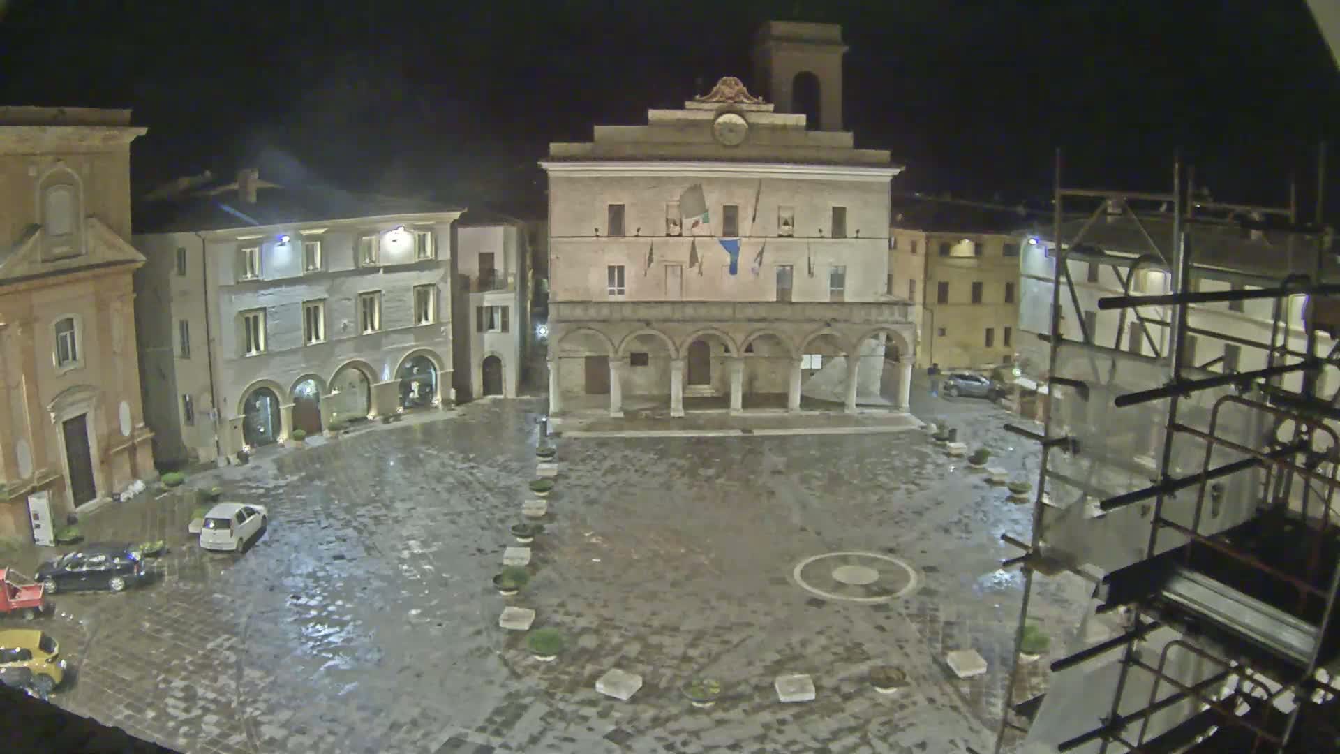 A wet, nighttime town square is seen, centered around a large, arched building with a clock tower, surrounded by other buildings and scaffolding.