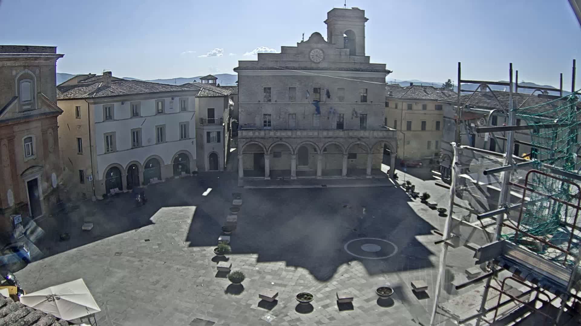 A large town square with a clock tower and surrounding buildings is bathed in sunlight.