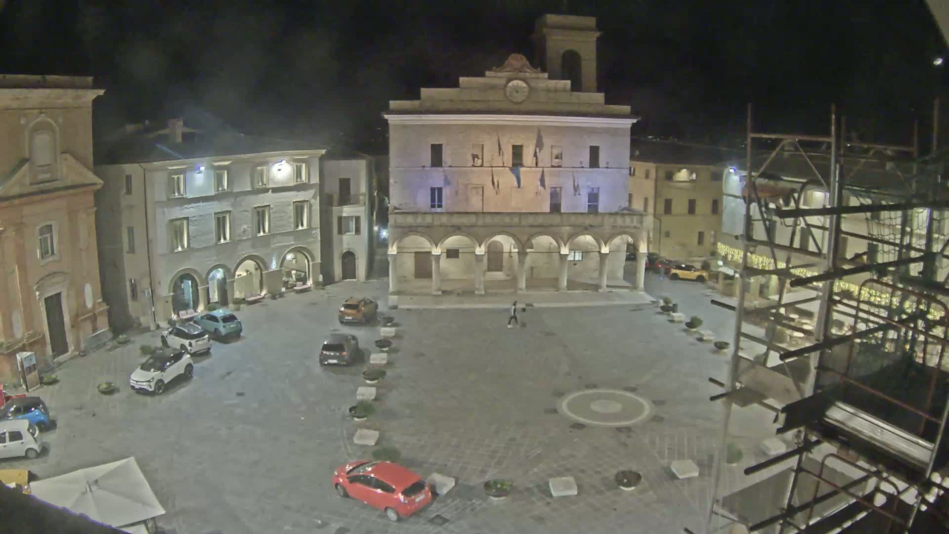 A nighttime view of a town square with several cars parked and a large building with arched entryways, under a dark sky.