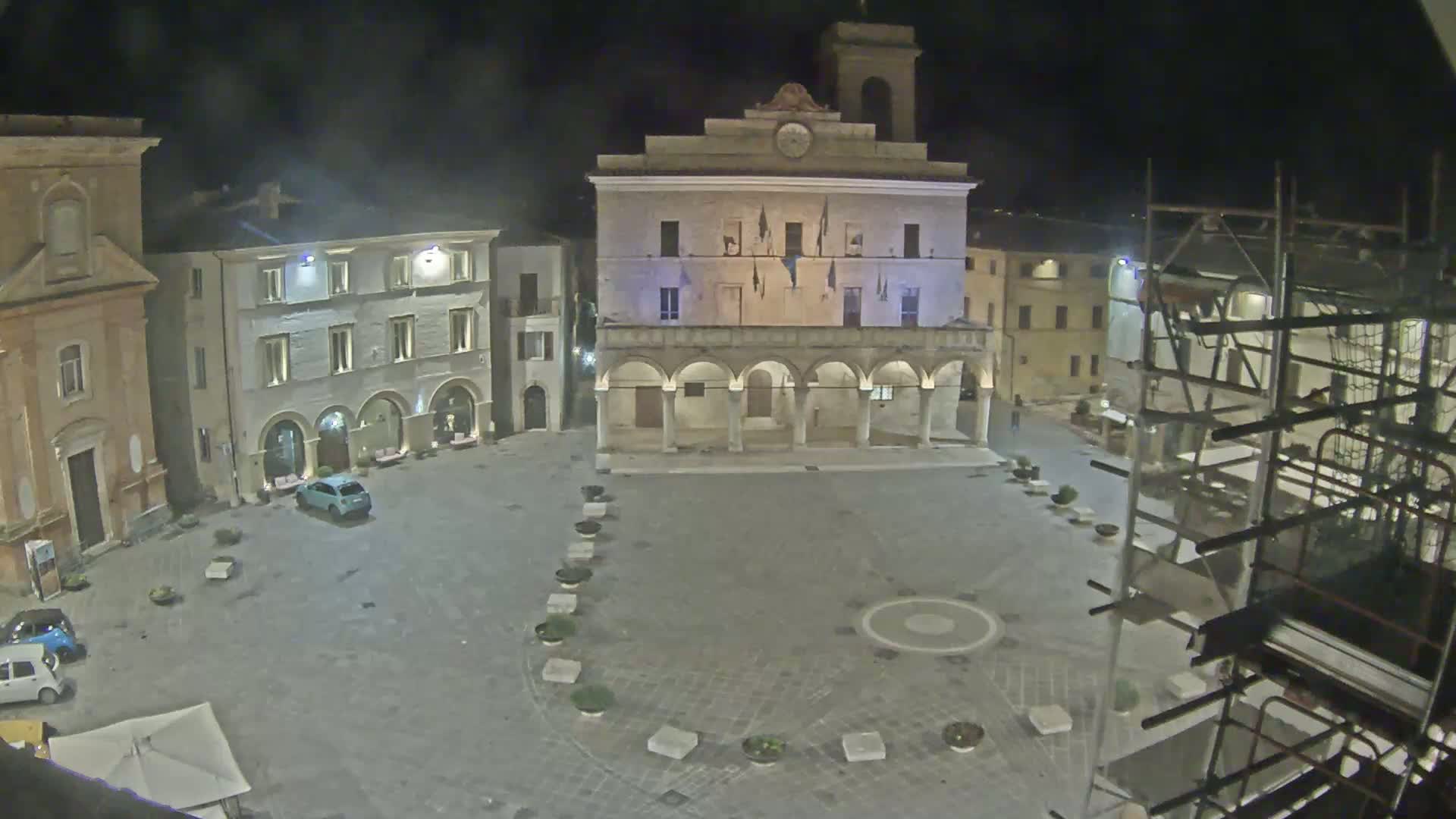 A nighttime view of a town square with several buildings, including a large building with arched entryways, a few cars, and scaffolding.