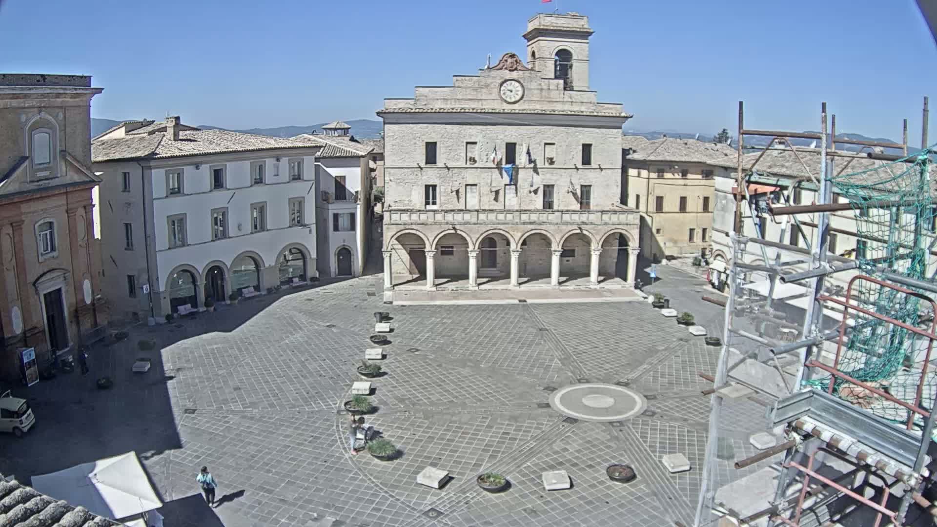 A large, paved town square on a sunny day is centered on a grand building with arched entryways, surrounded by other buildings, and partially obscured by scaffolding on one side.