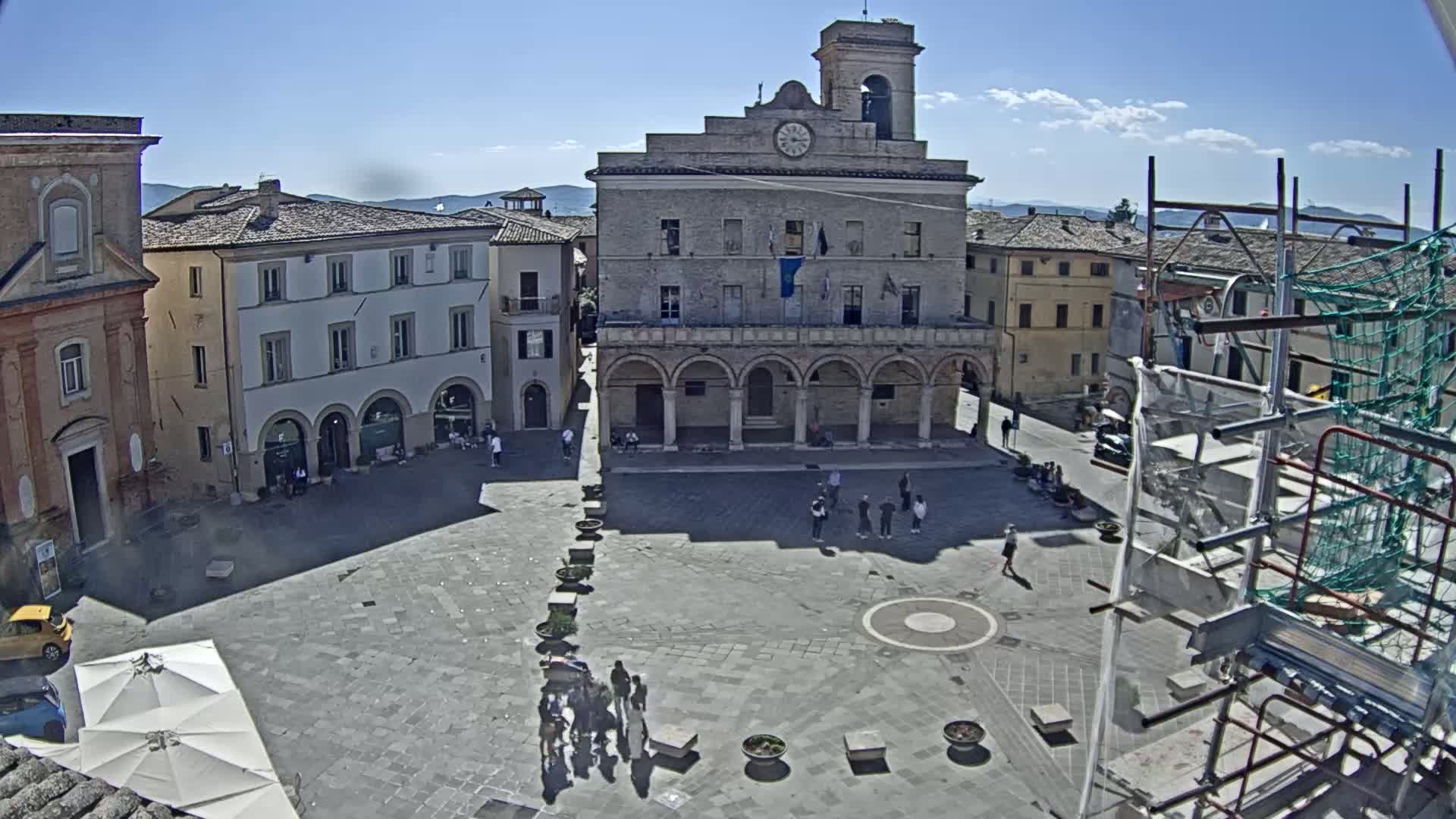 A sunny town square with a large stone building, surrounded by other buildings and a few people.