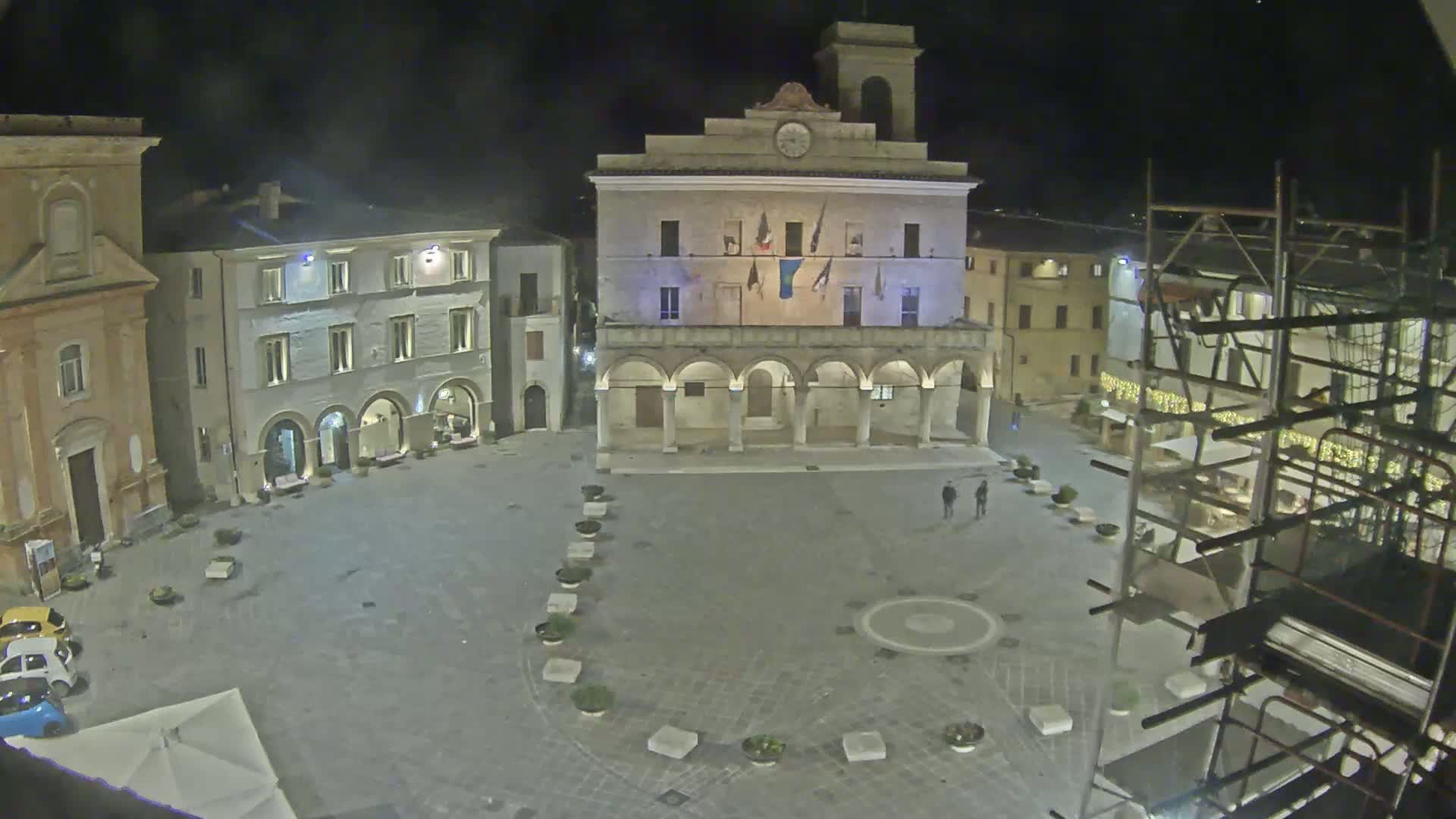 A nighttime view of a town square with several buildings, including a large building with arched entryways, under a clear, dark sky.