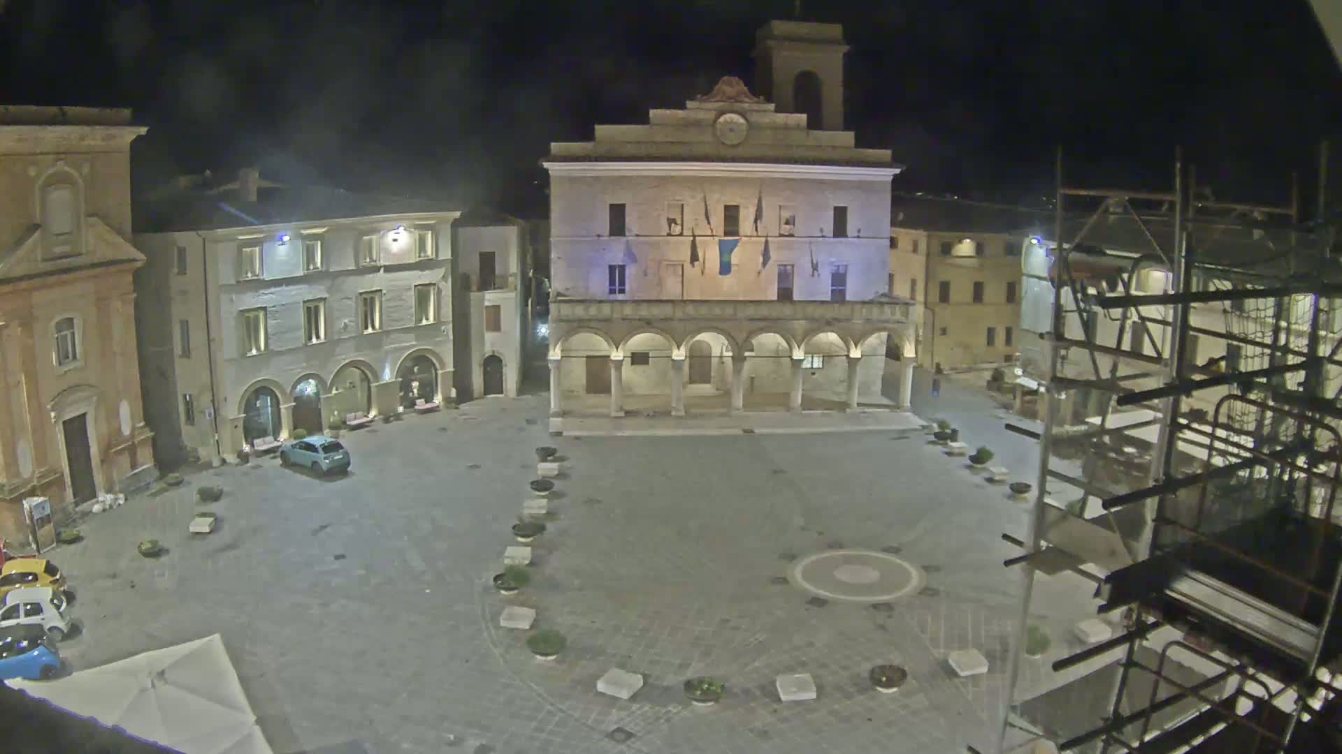 An empty town square at night is illuminated, showing a large building with arched entryways, other buildings surrounding it, a few cars parked, and scaffolding on one side.