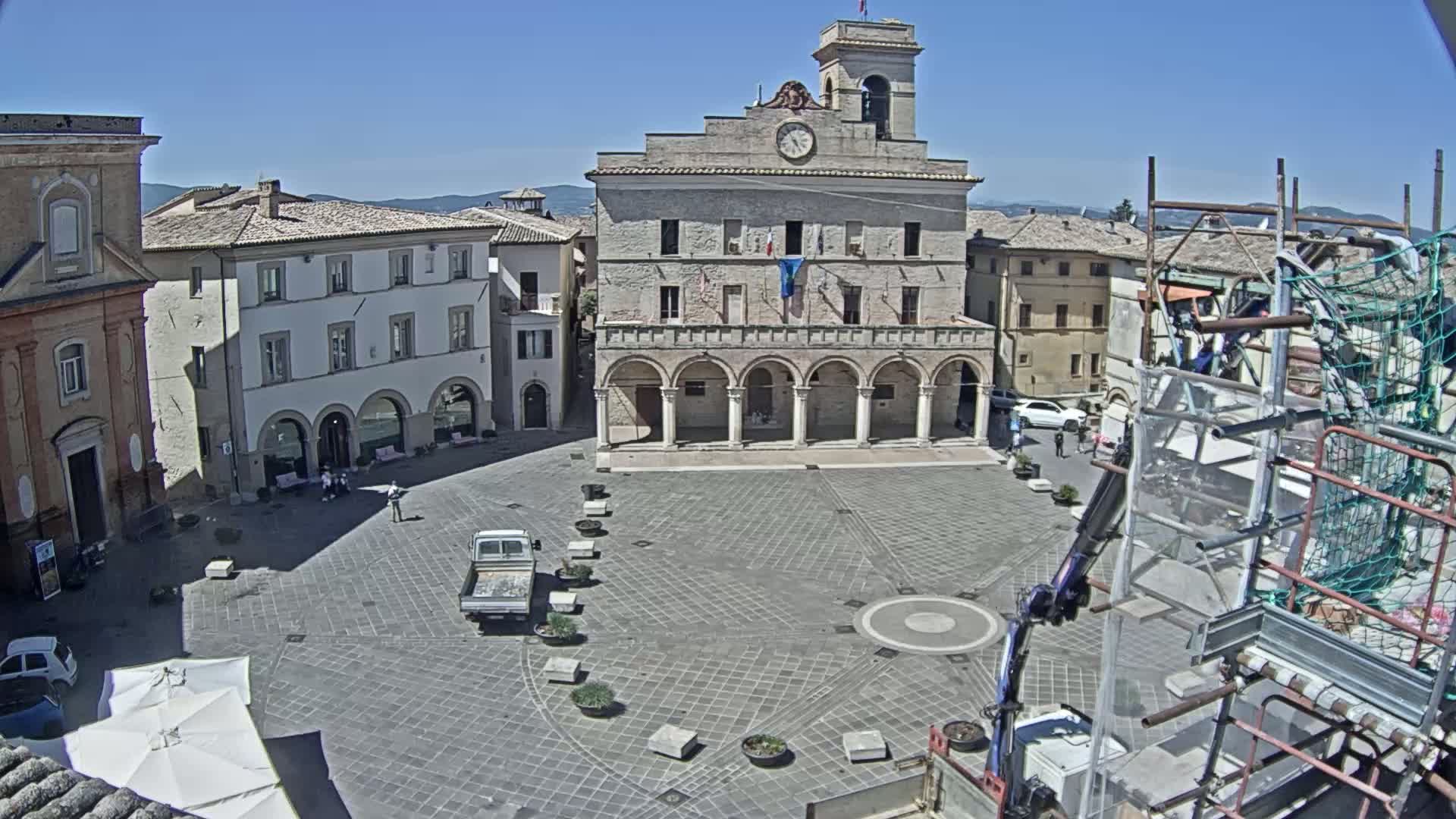 A large town square with a clock tower and surrounding buildings is seen under sunny skies.