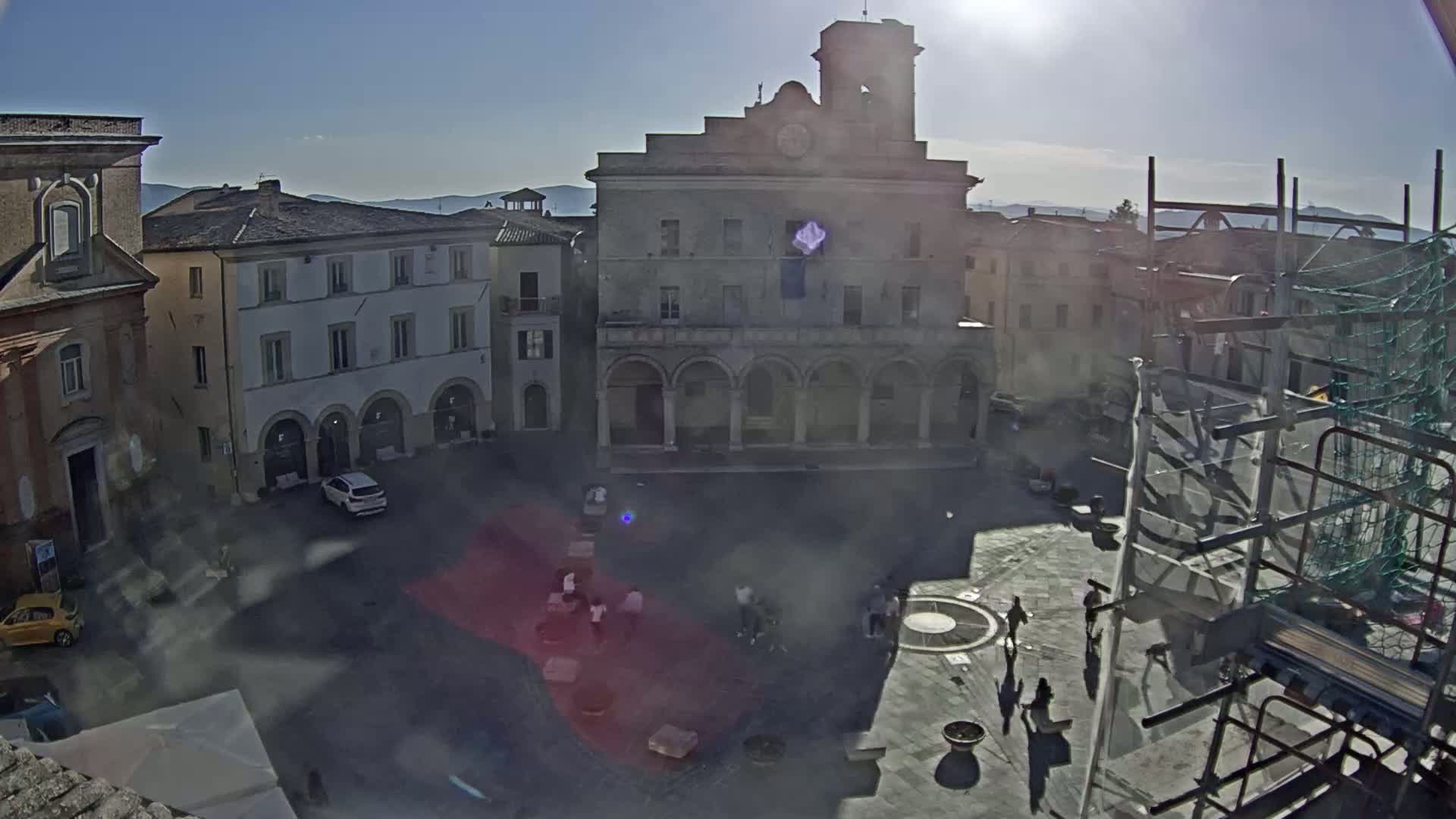 A sunny, high-angle view shows a town square with several buildings, a few cars, and people walking around.