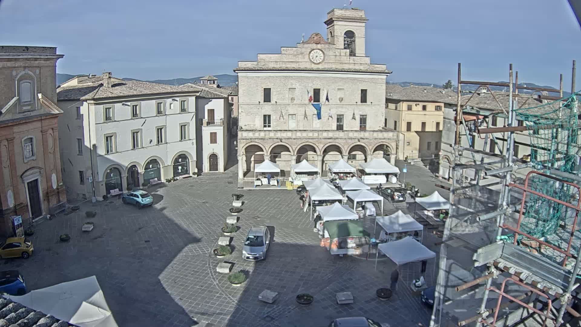 A sunny day reveals a town square with a market set up in front of a large, arched building, surrounded by other buildings, some under construction.