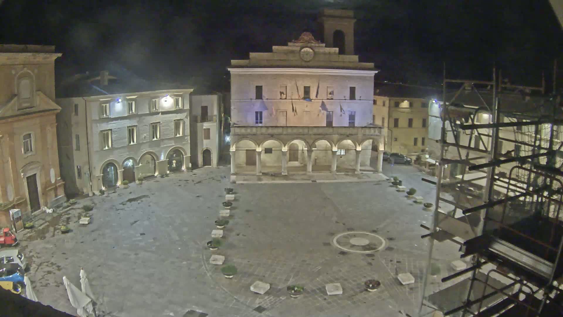 A town square at night, wet from recent rain, is surrounded by buildings including a large, columned structure, and scaffolding is visible to the right.