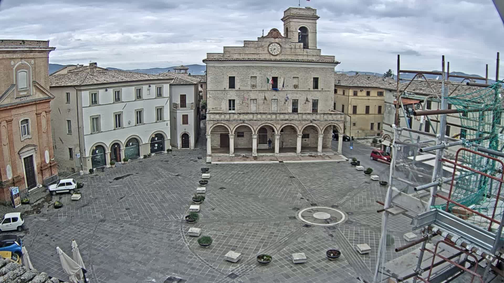 A town square with several buildings, including a large stone building with arched entryways, under an overcast sky.