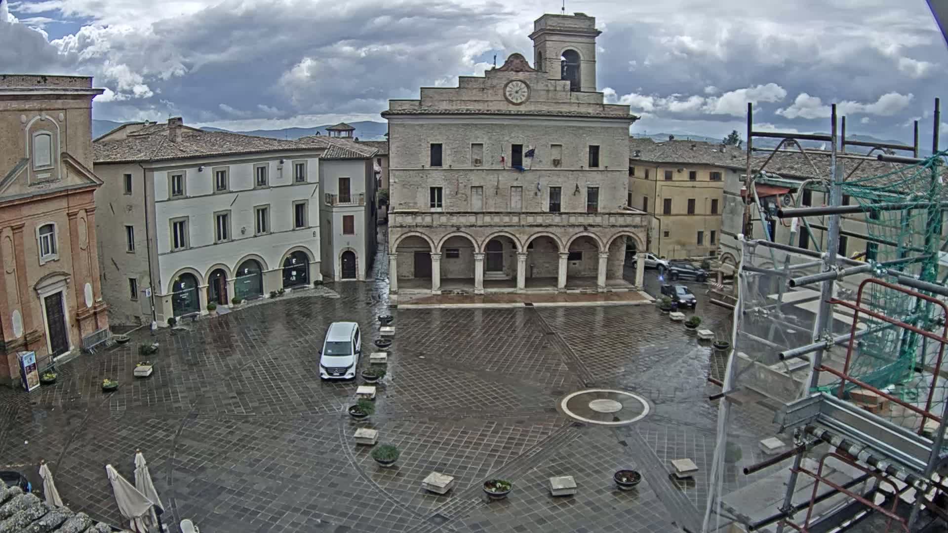 A wet town square is centered around a large building with arched entryways, surrounded by other buildings under a cloudy sky.