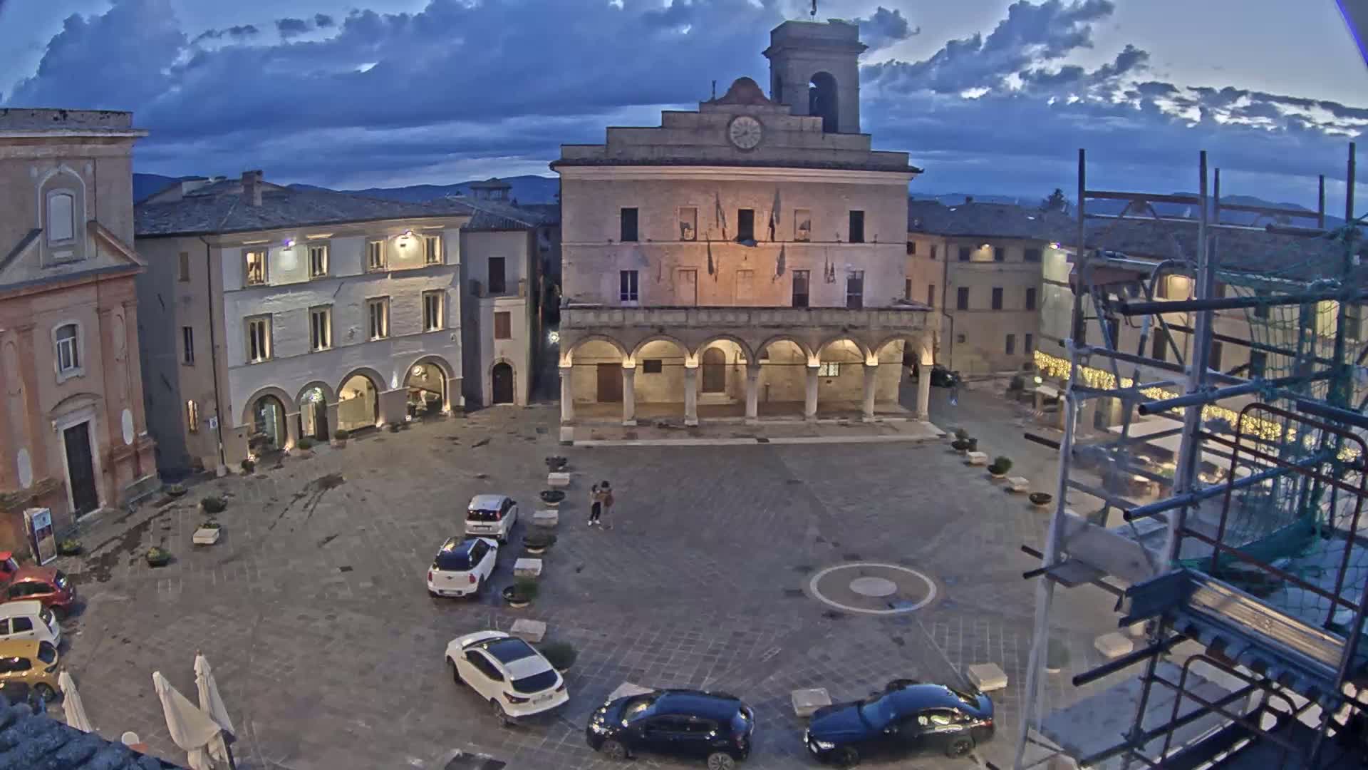 A town square under a cloudy twilight sky is populated by several cars and a few pedestrians near a large building with arched entryways.