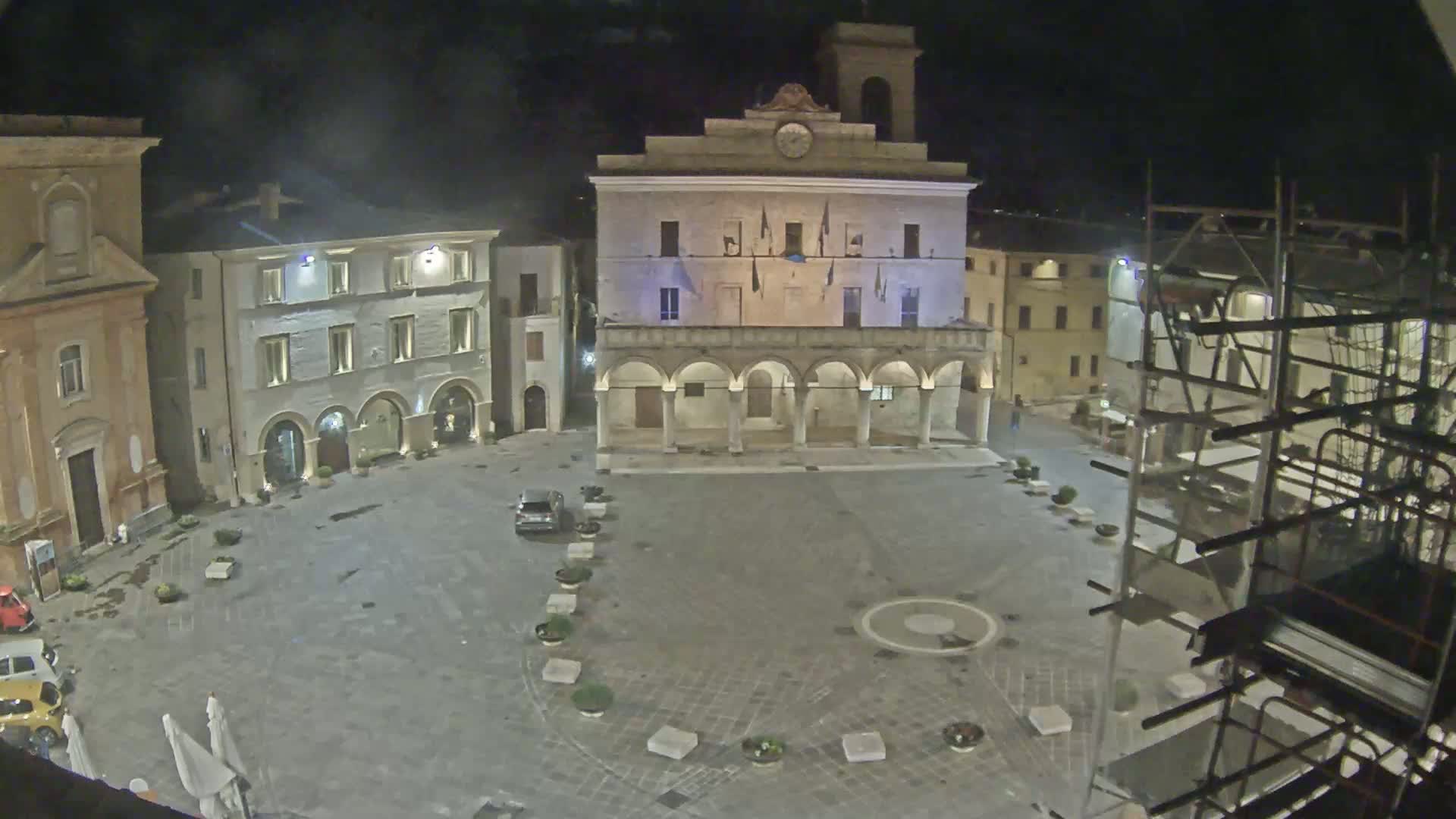 A nighttime view of a town square with several buildings surrounding it, one of which is a large structure with arched walkways, under dark, clear skies.