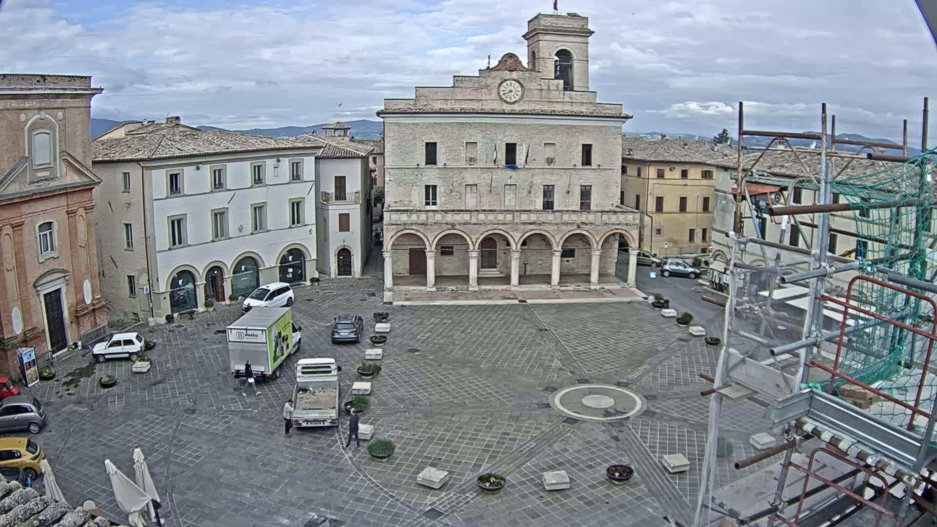 A large, stone plaza with a clock tower and several buildings surrounding it under an overcast sky; construction scaffolding is visible in the lower right corner.