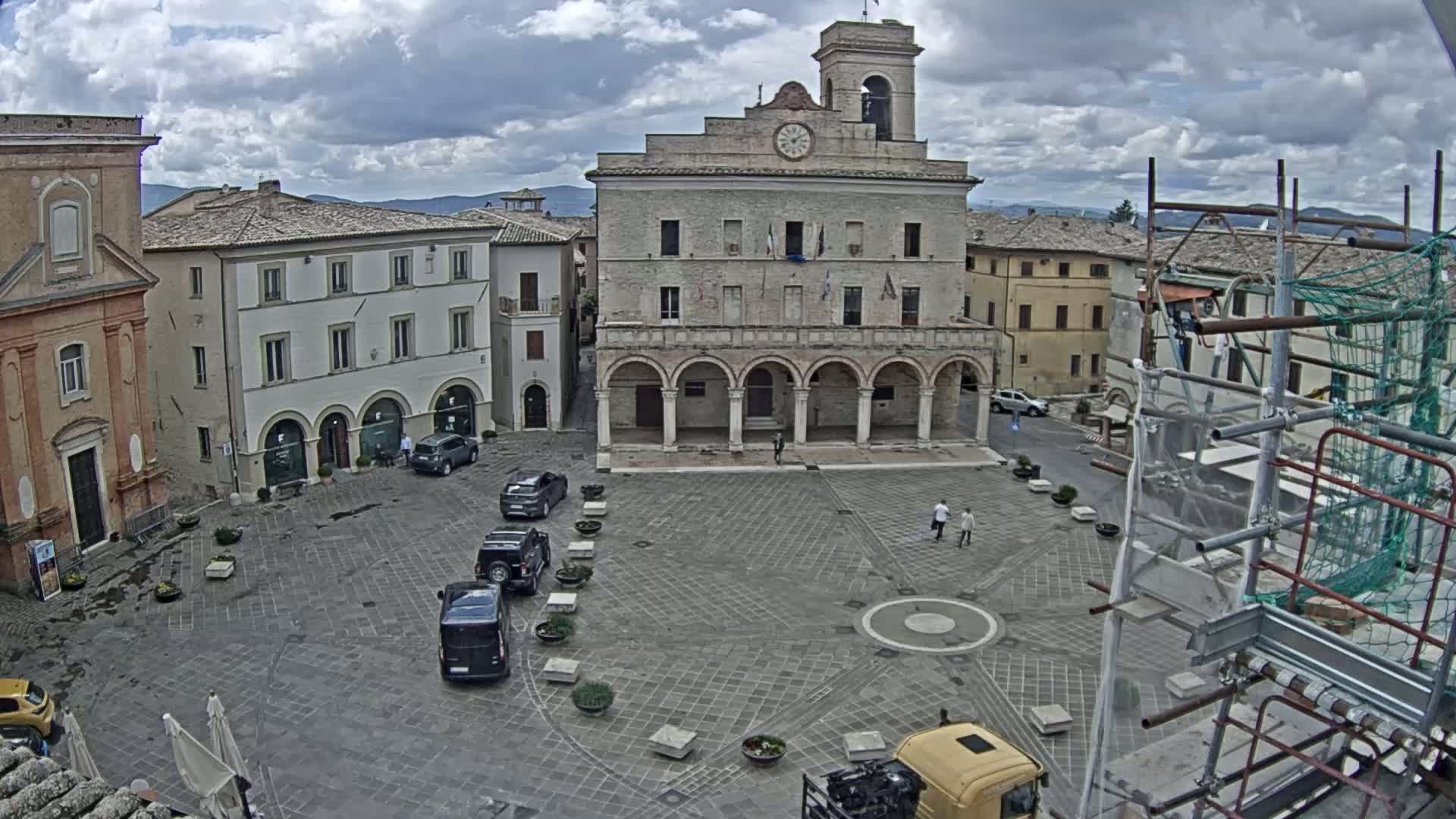 A town square with a large stone building, several cars parked, and scaffolding in the lower right corner under a cloudy sky.