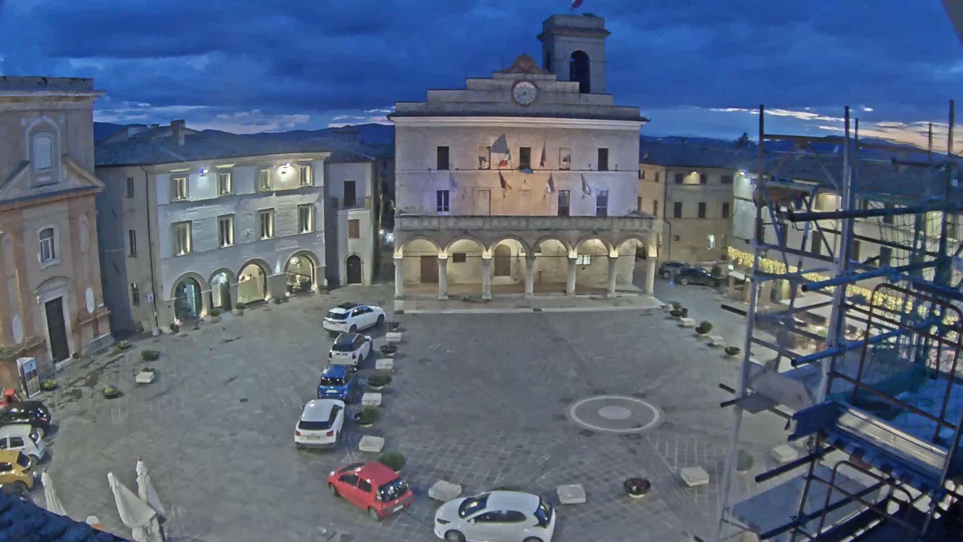 A town square at dusk, under a partly cloudy sky, contains several parked cars and is bordered by historic buildings, one of which is a large civic building with a colonnade.