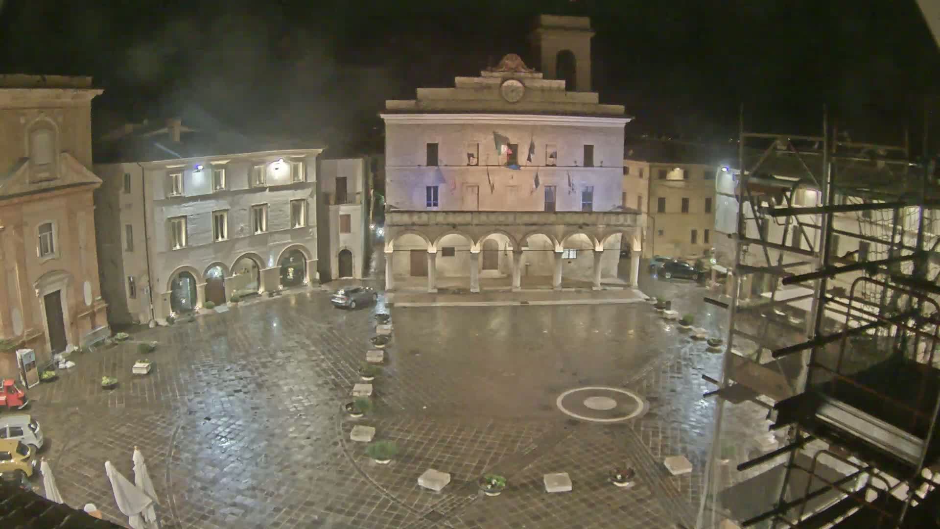 A wet, cobblestone town square at night is surrounded by historic buildings, one of which has an arched colonnade, with scaffolding visible in the corner.