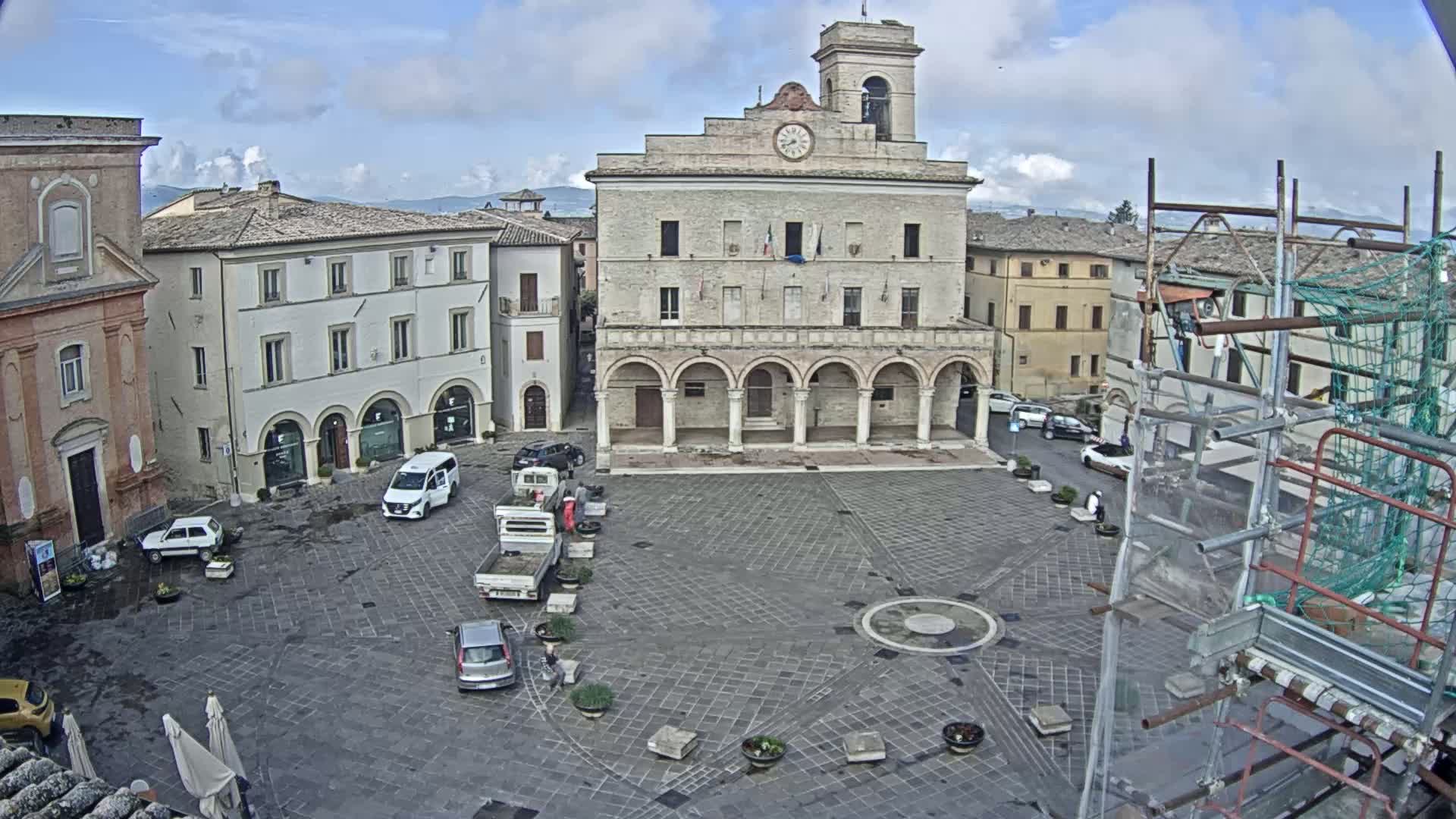 A town square with several parked cars, construction scaffolding, and a large building with arched entryways under partly cloudy skies.
