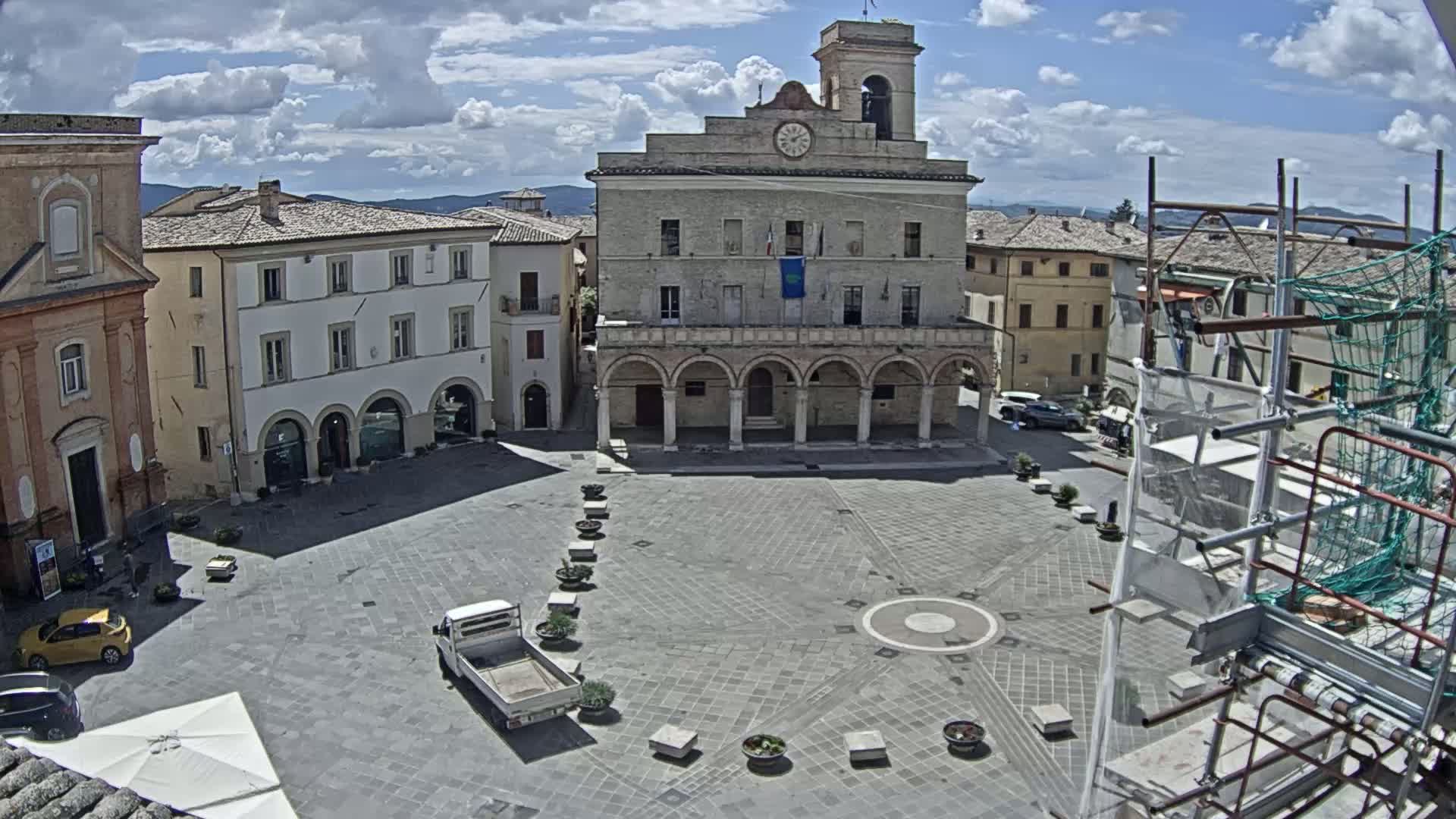 A partly cloudy day reveals a town square with a large building featuring arched entryways, surrounded by other buildings under a partly cloudy sky.