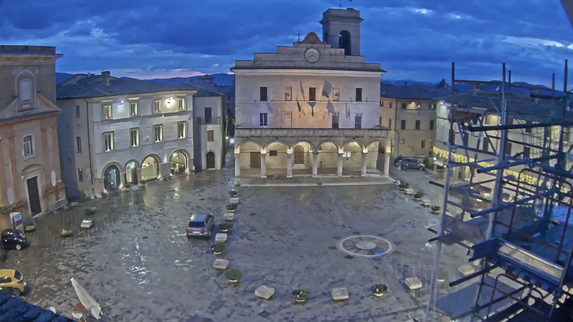 A wet town square at twilight is centered around a large building with arched entryways, surrounded by other buildings and scaffolding, under a partly cloudy sky.