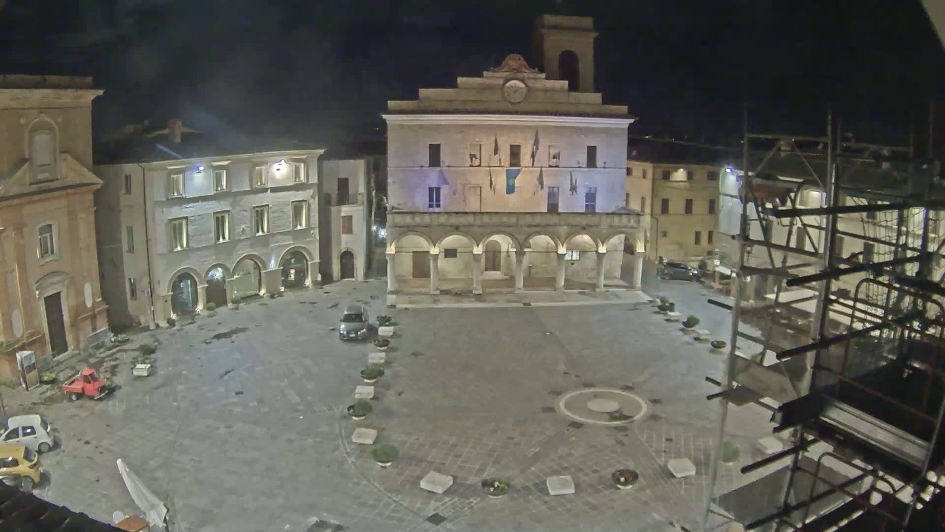 A town square at night, illuminated, showing several buildings including a large stone building with arched entryways, and a few cars parked in the square.