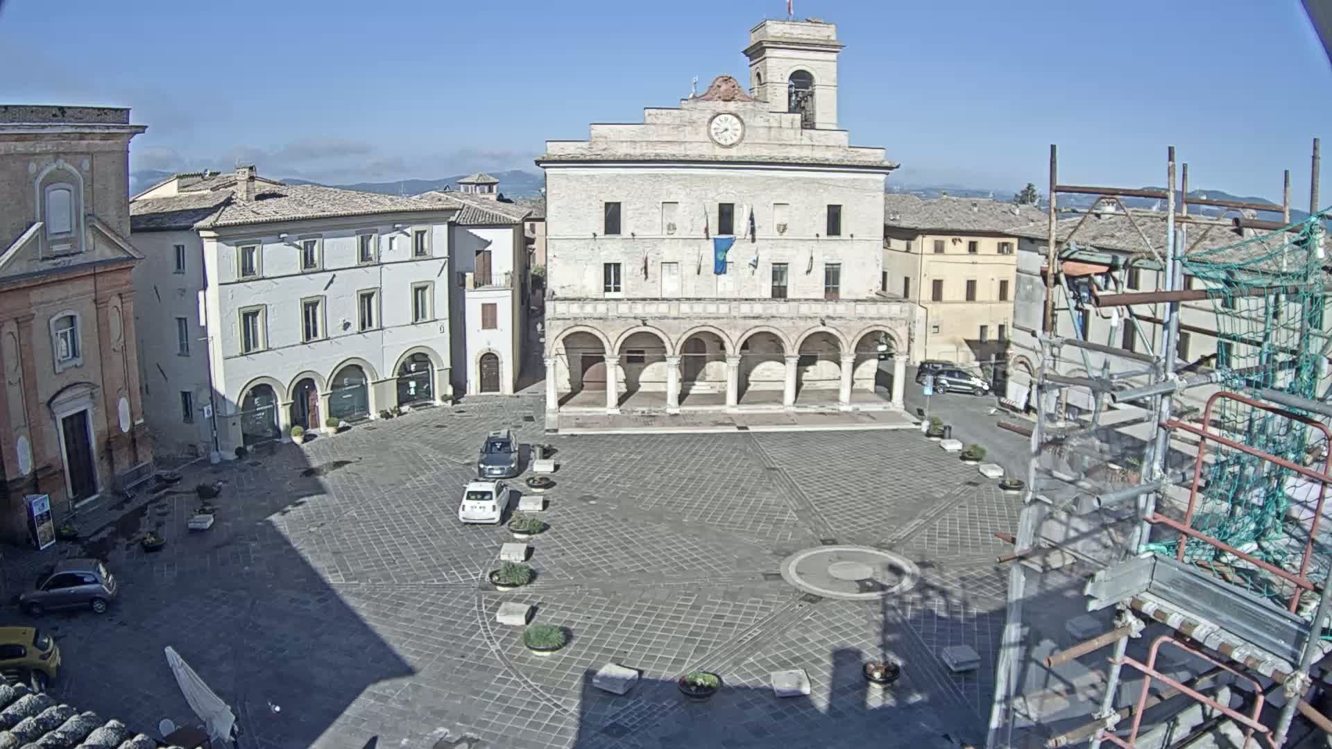 A large, paved town square is centered around a building with arched walkways under a clear, sunny sky, with a few cars and scaffolding visible on the perimeter.