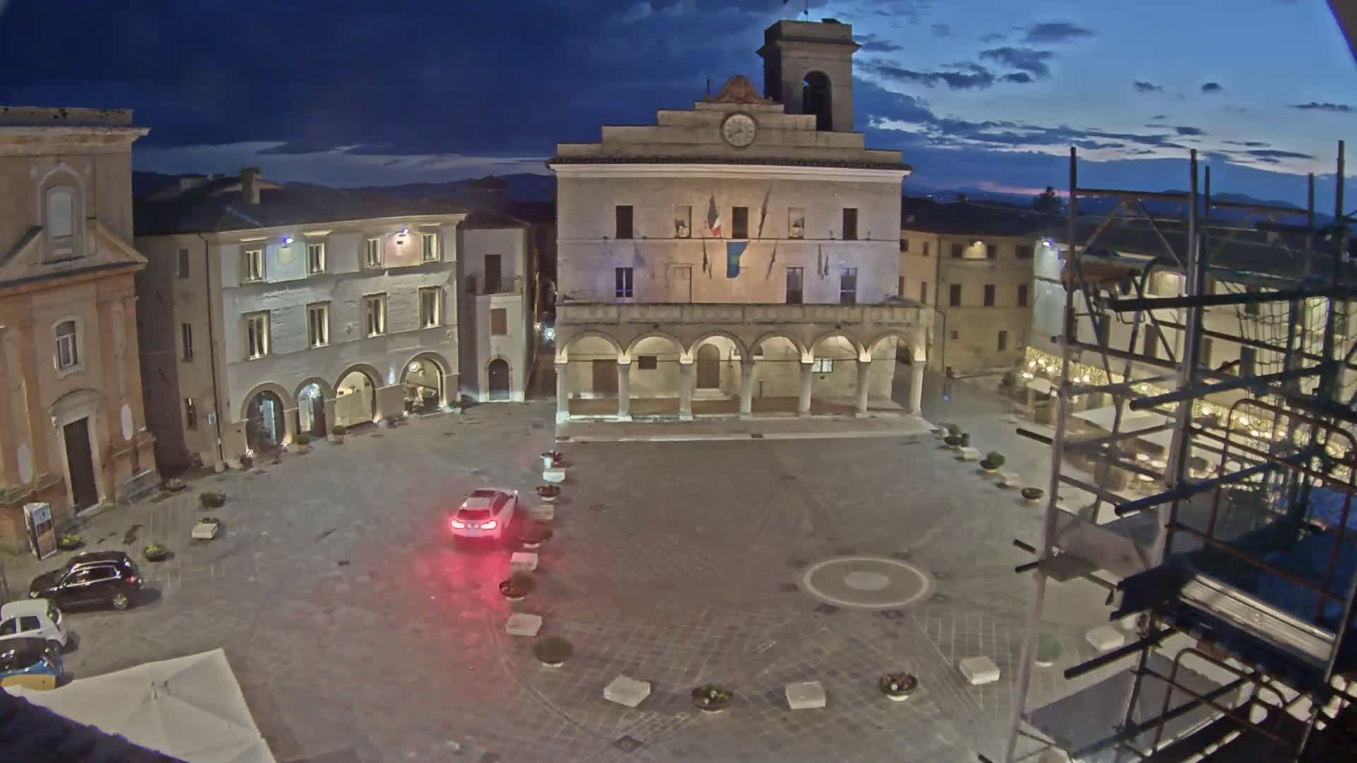 A town square at twilight is illuminated, featuring a large building with arched entryways, several smaller buildings, a few cars, and scaffolding near a restaurant.