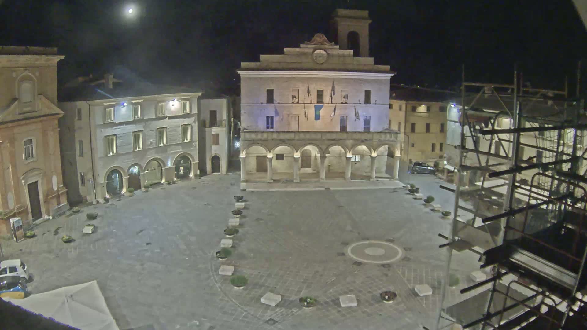 A moonlit town square at night features a large building with arches, surrounded by other buildings, some with scaffolding.