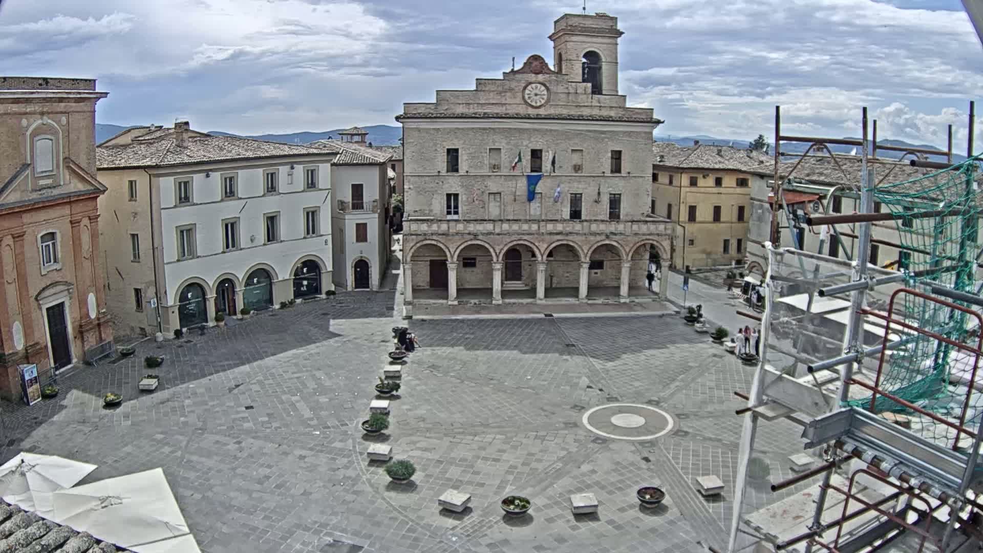 A town square, under a partly cloudy sky, features a large stone building with arched entryways, surrounded by other buildings.